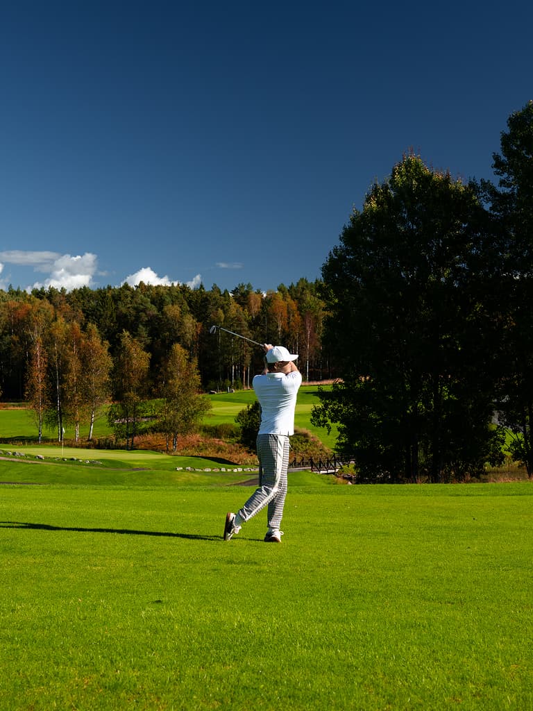 Golfer in weißem Shirt und Cap schwingt den Schläger auf einem grünen Fairway, mit herbstlichen Bäumen und tiefblauem Himmel.