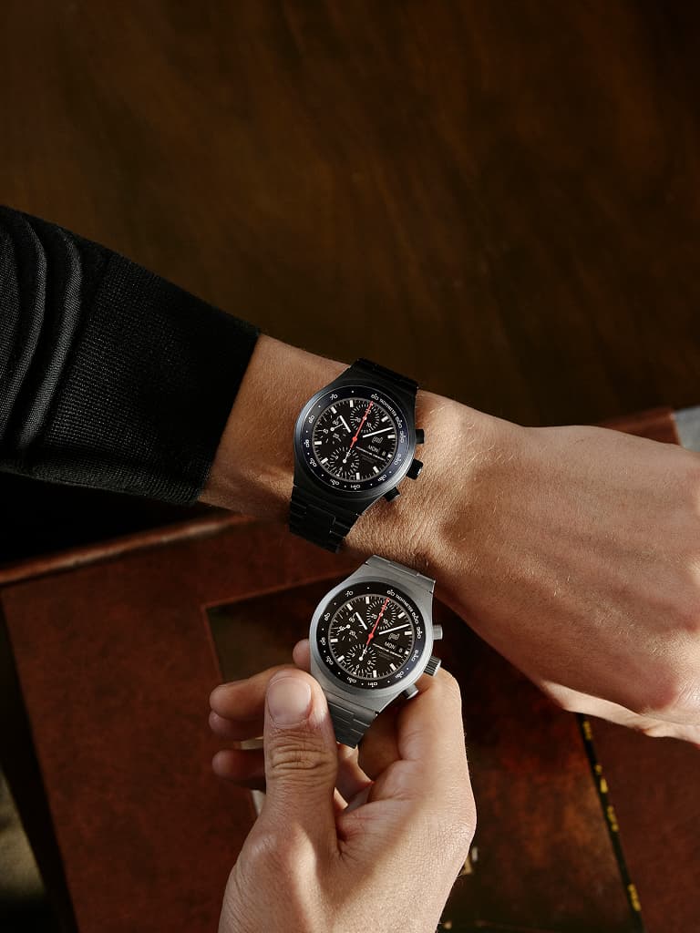 Dos relojes de pulsera cronógrafo con esfera negra, uno puesto en la muñeca y otro sostenido en la mano, sobre una mesa de madera.