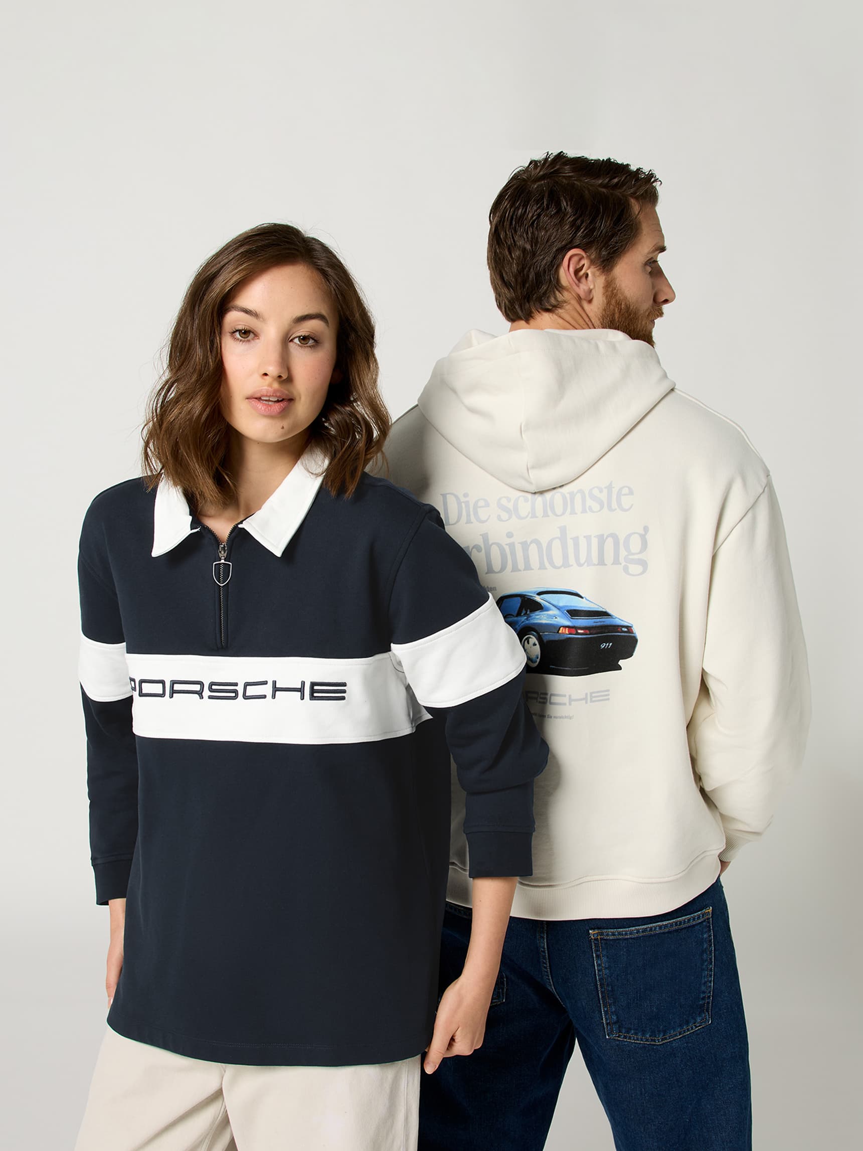 Twee modellen in Porsche-kleding: vrouw in marineblauw top met witte streep, man in crèmekleurige hoodie met autografiek.