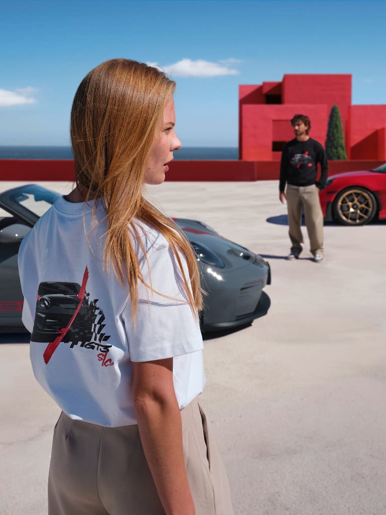 Woman wearing a white T-shirt with back print, standing on a sunny seaside terrace in front of a gray and red sports car.