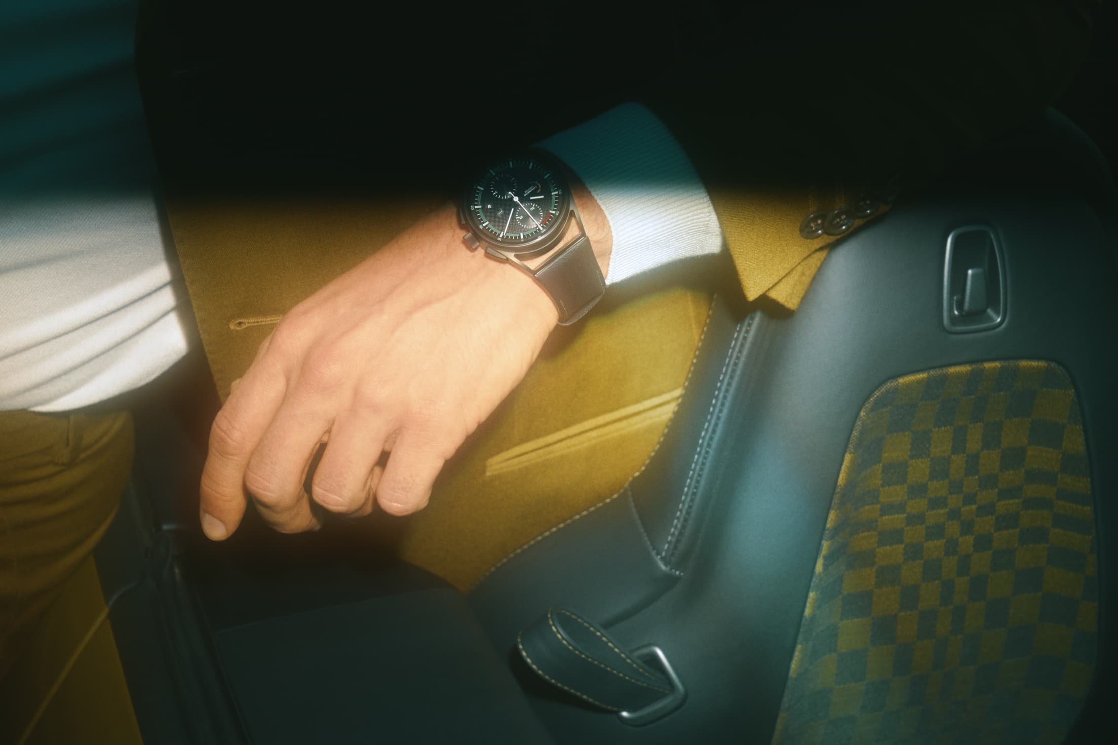 Détail d'un homme portant un costume olive et assis dans une Porsche, il porte une montre Porsche Design.