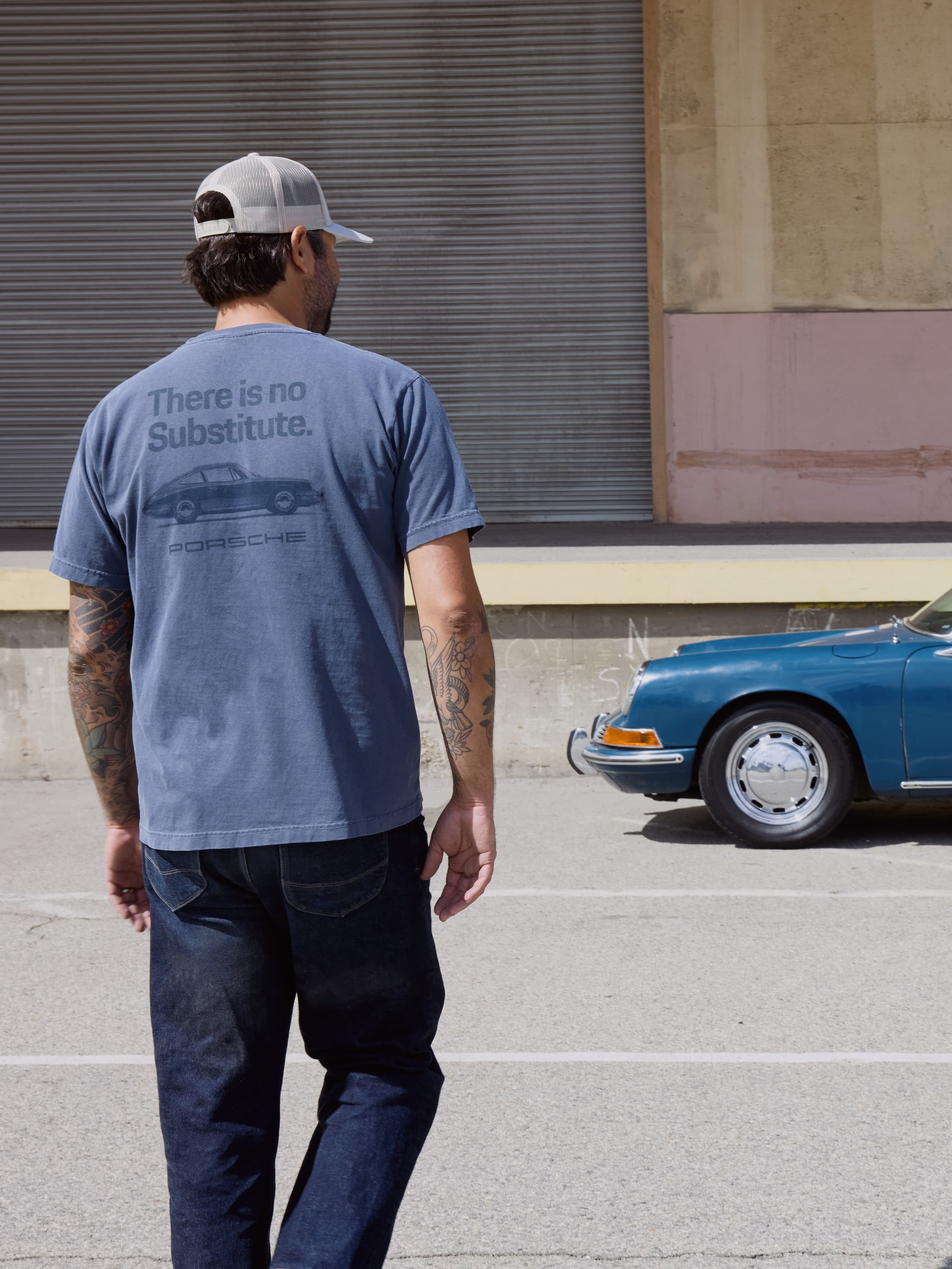 Male Model wearing Porsche There is no Substitute T-Shirt walking towards a blue Porsche.