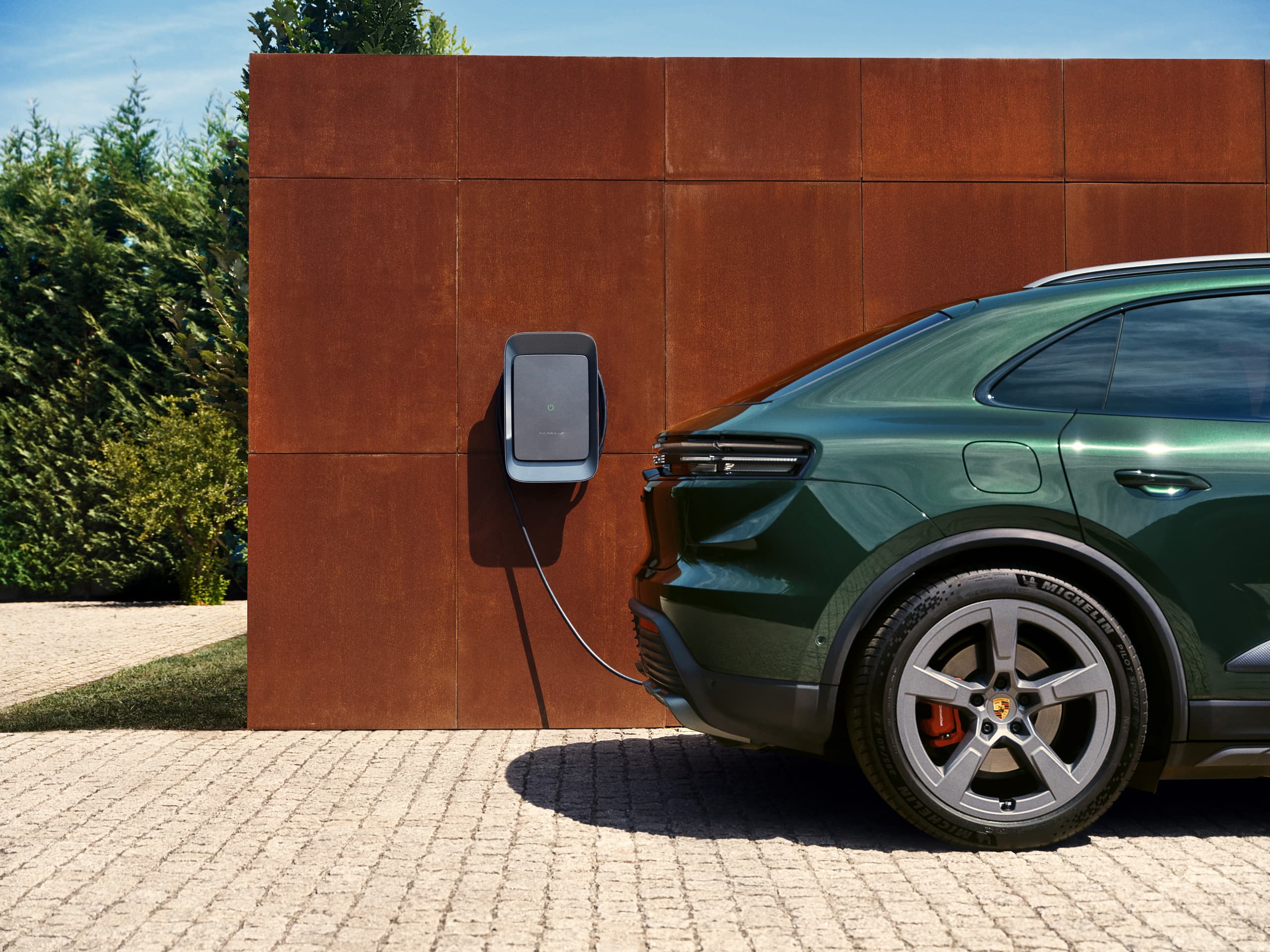 the rear part of a dark green Porsche Macan on a paved floor in front of a rust-red wall with a Porsche Wallbox, plants and blue sky in the background
