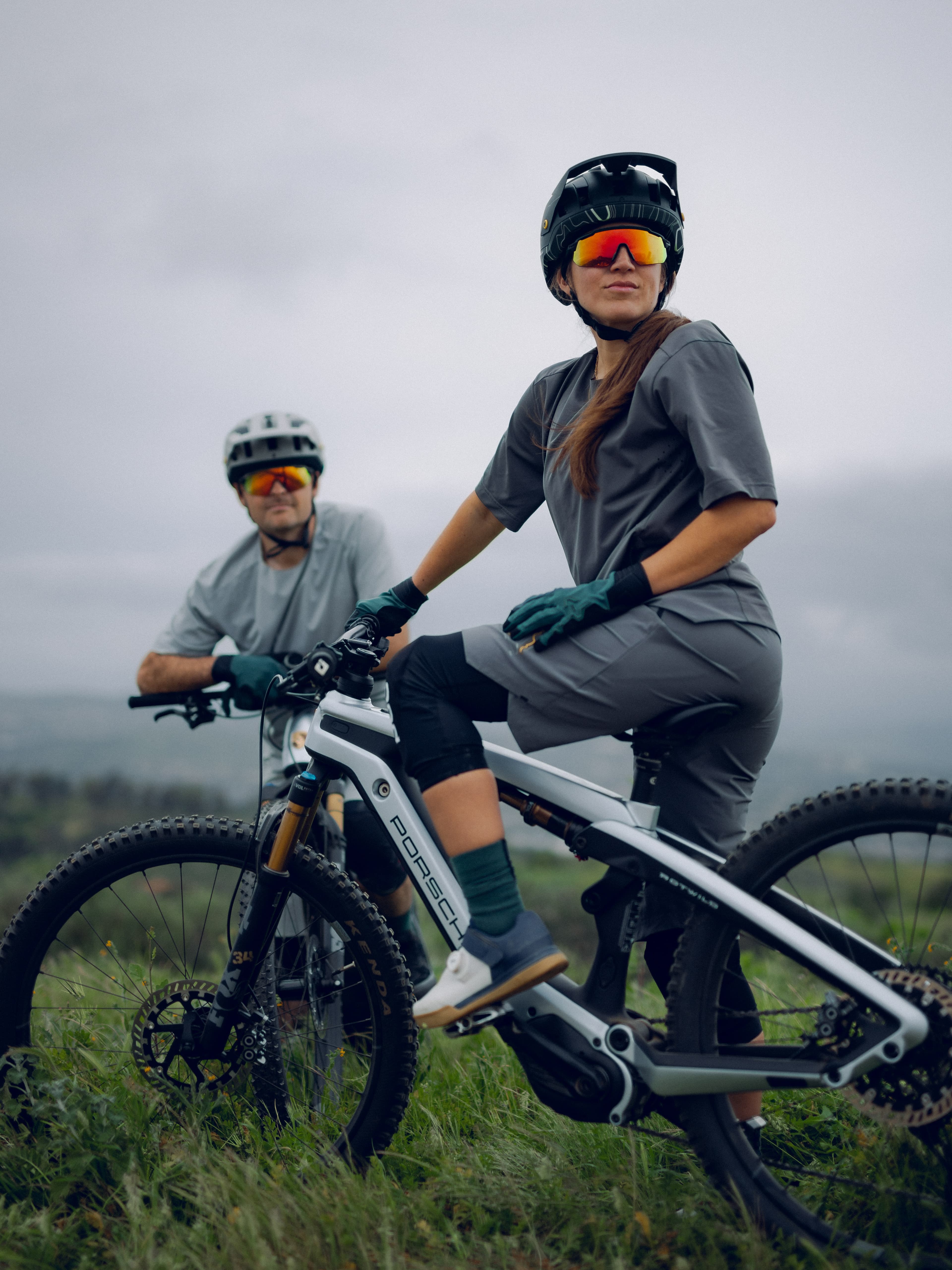 Deux cyclistes en tenue grise sur des vélos Porsche, portant des casques et des lunettes de soleil, dans un paysage herbeux sous un ciel nuageux.