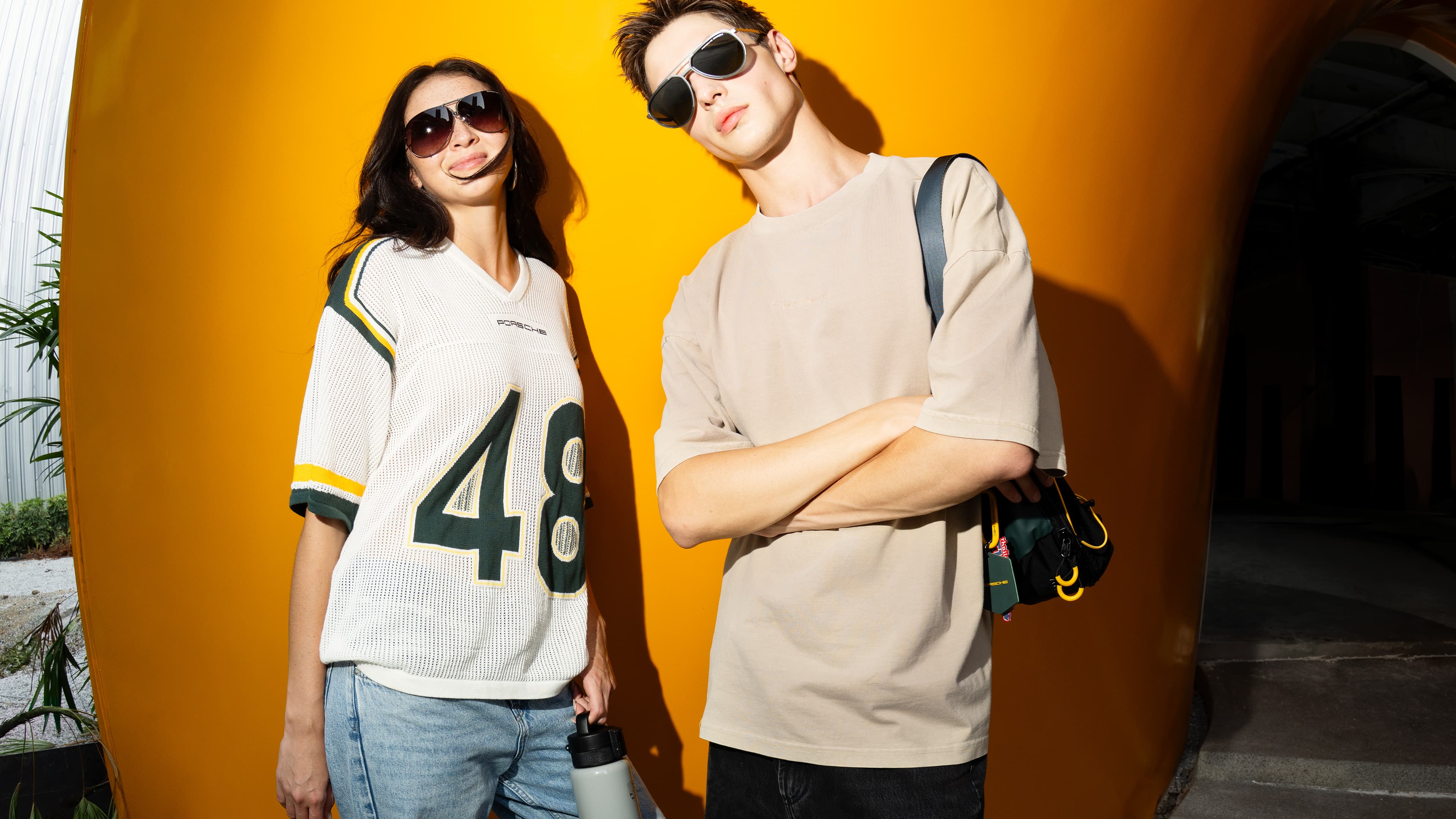 Two models wearing sunglasses in front of a yellow wall, wearing a Porsche shirt and a beige T-shirt, with a bag and a water bottle from the new AHEAD collection.