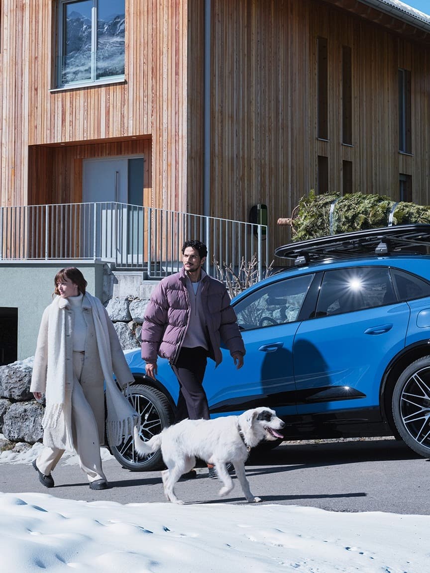 Couple walking dog by blue SUV with tree on roof, in front of wooden house, snowy ground.