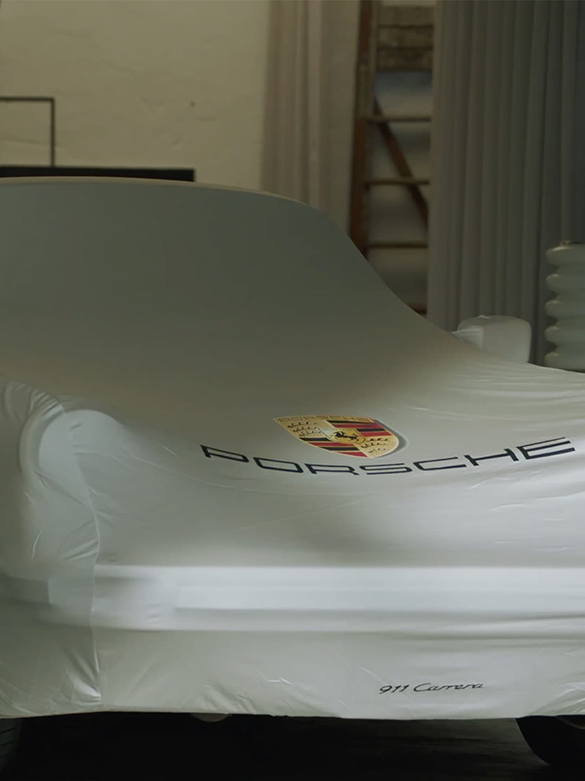 Porsche 911 Carrera under a white cover with logo, in a dimly lit garage setting.
