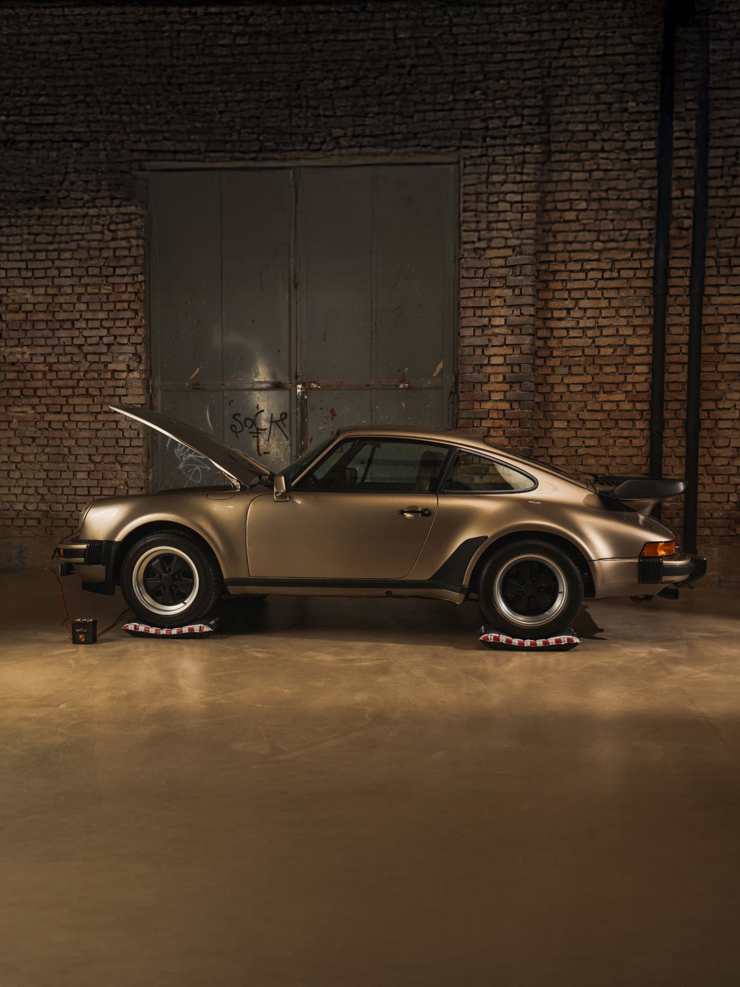 Gold Porsche 911 with open hood in a dimly lit garage, parked on tire cushions, brick wall background.