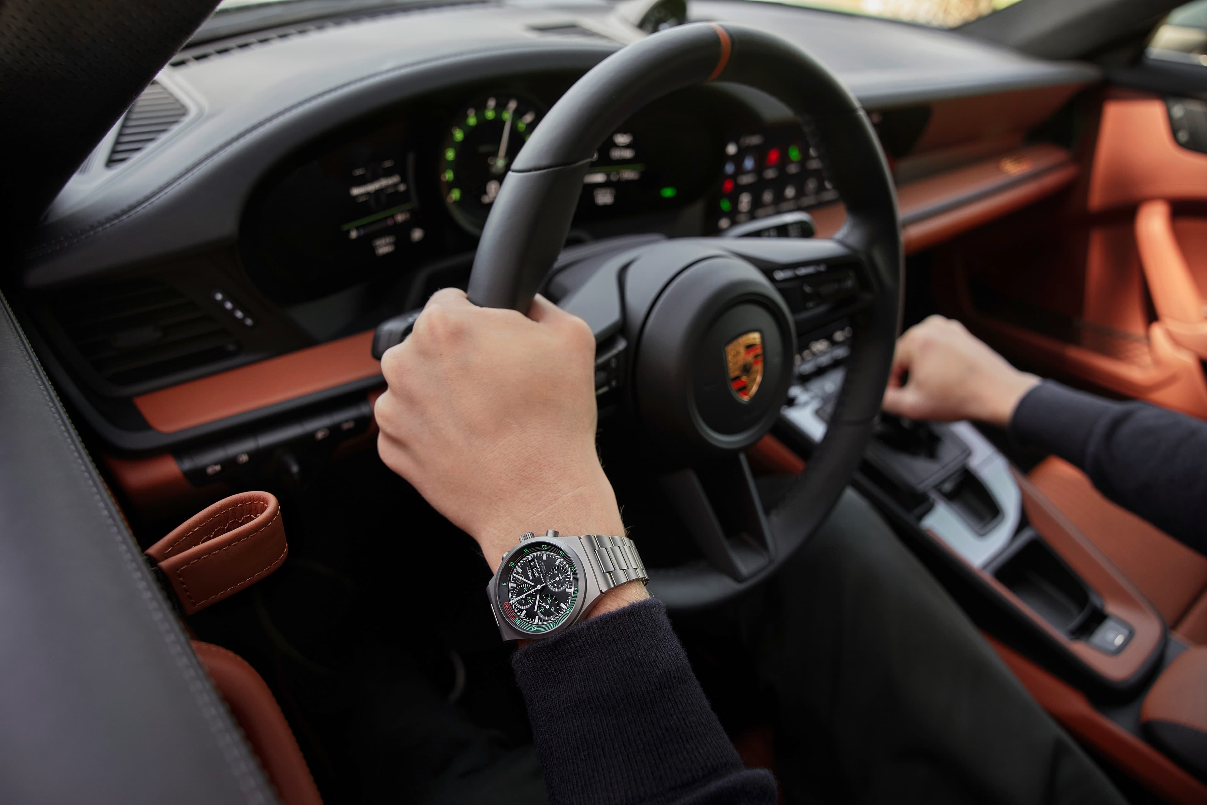 Hand am Porsche-Lenkrad, Edelstahl-Chronograph am Handgelenk, Innenraum in Schwarz und Cognac-Leder.