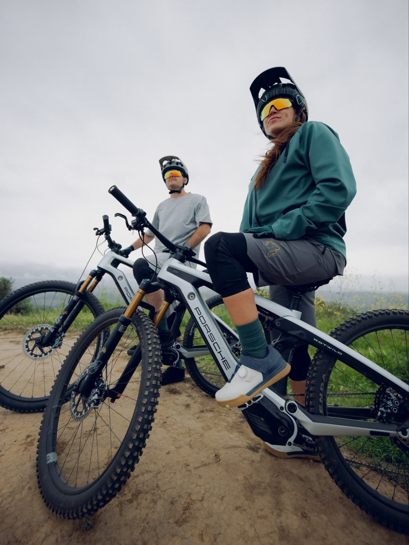 Two mountain bikers wearing helmets and mirrored sunglasses on gray e-bikes, taking a break on a dirt path under an overcast sky.