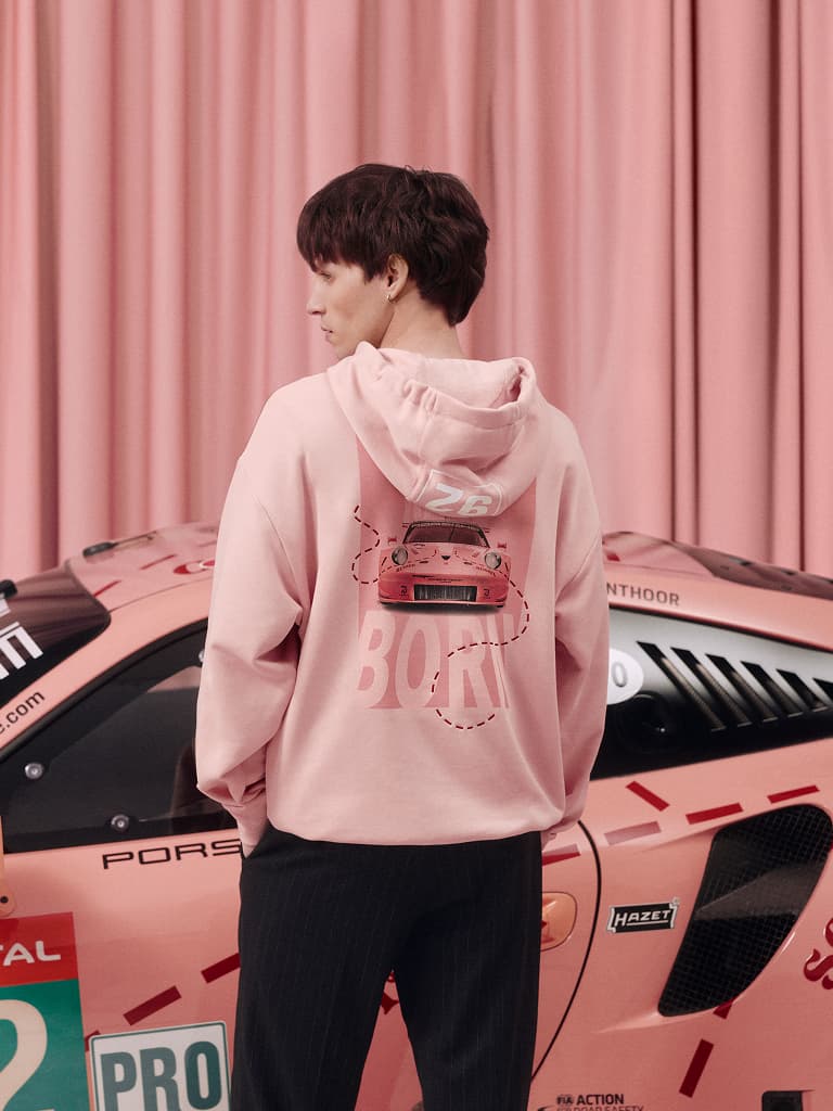 Personne en sweat à capuche rose avec graphique de voiture se tient à côté d'une Porsche de course rose, fond de rideau rose.