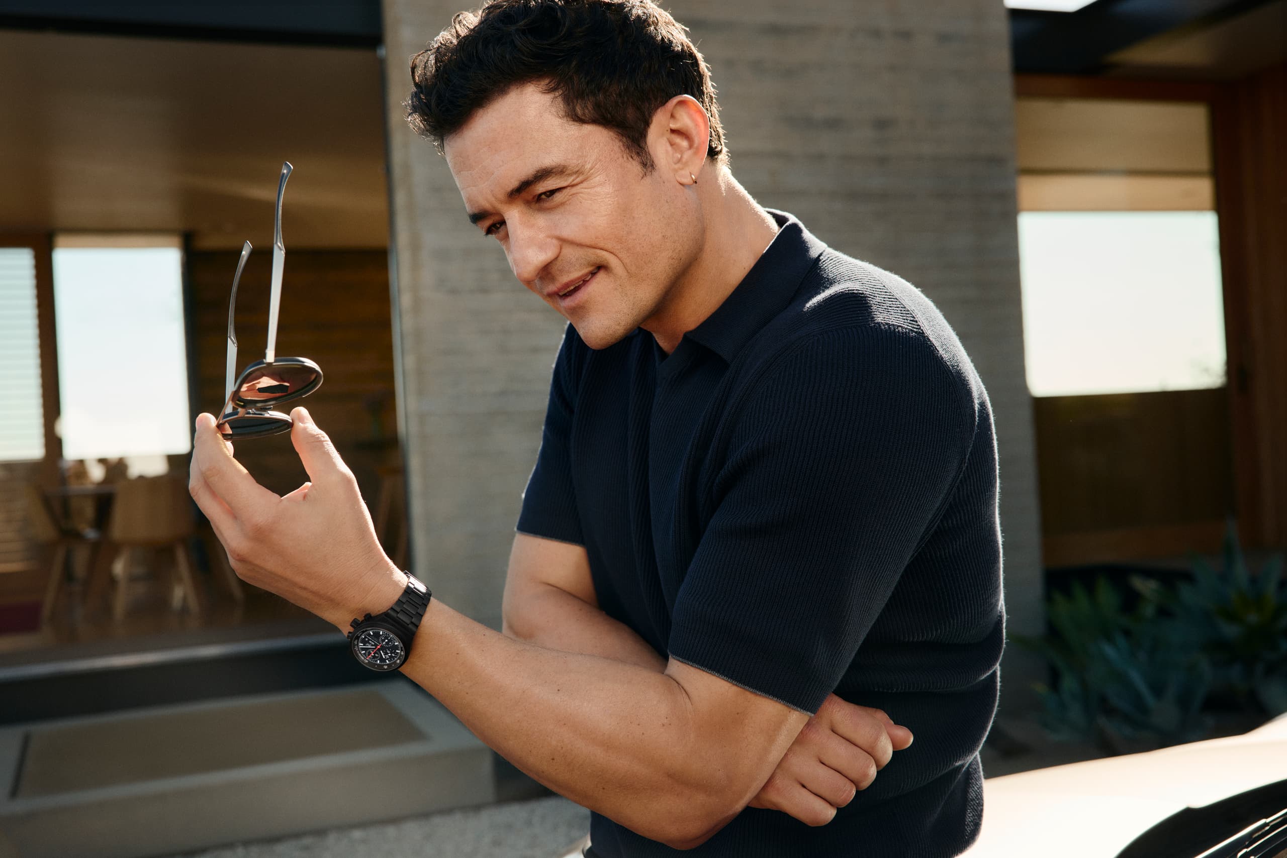 Orlando Bloom ma na nadgarstku zegarek Chronograph 1 – All Black Numbered Edition, stoi przed domem i uśmiecha się.