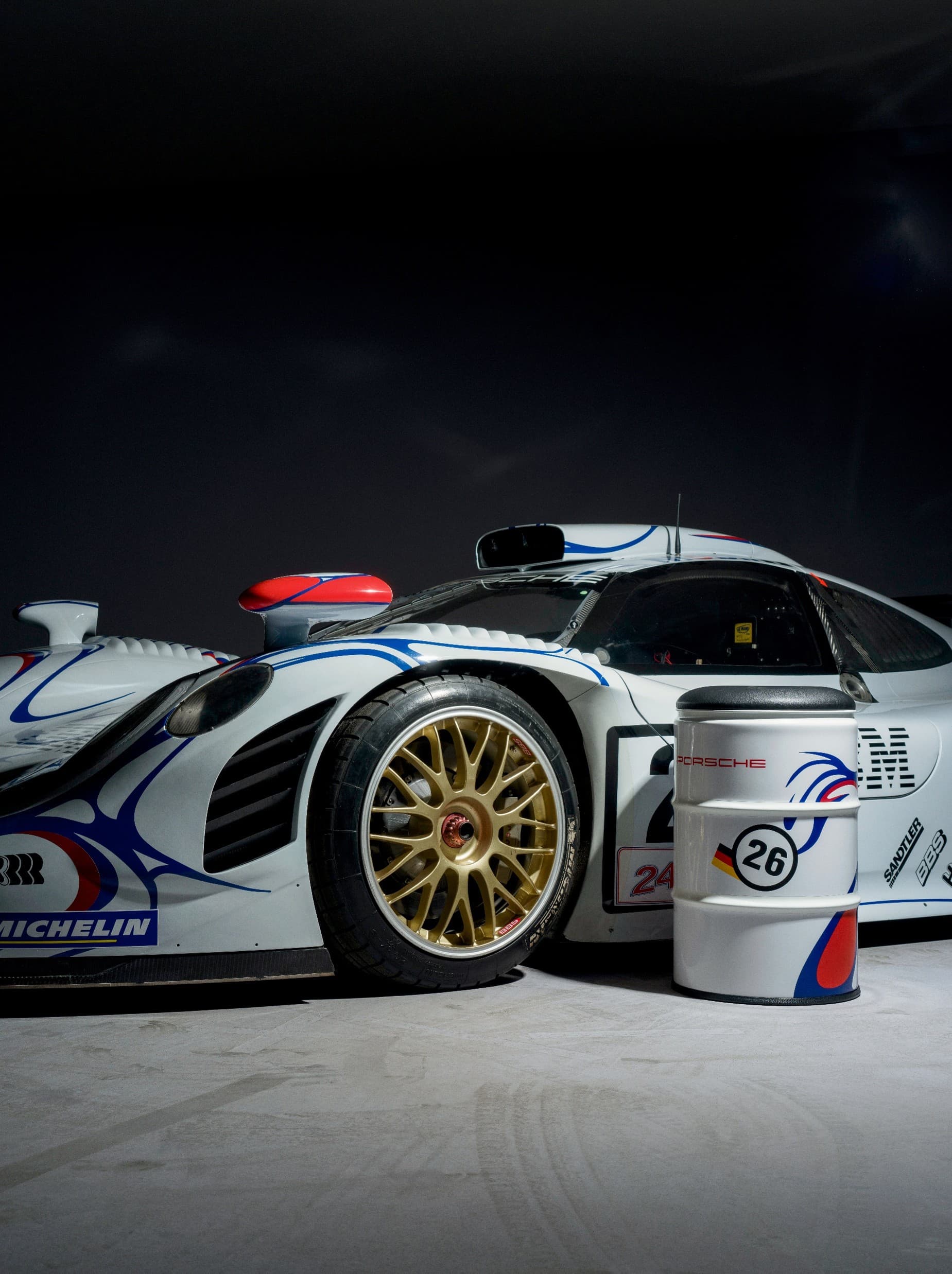Coche de carreras Porsche blanco con ruedas doradas, acentos rojos y azules, junto a un barril a juego con el número 26 sobre un fondo oscuro.