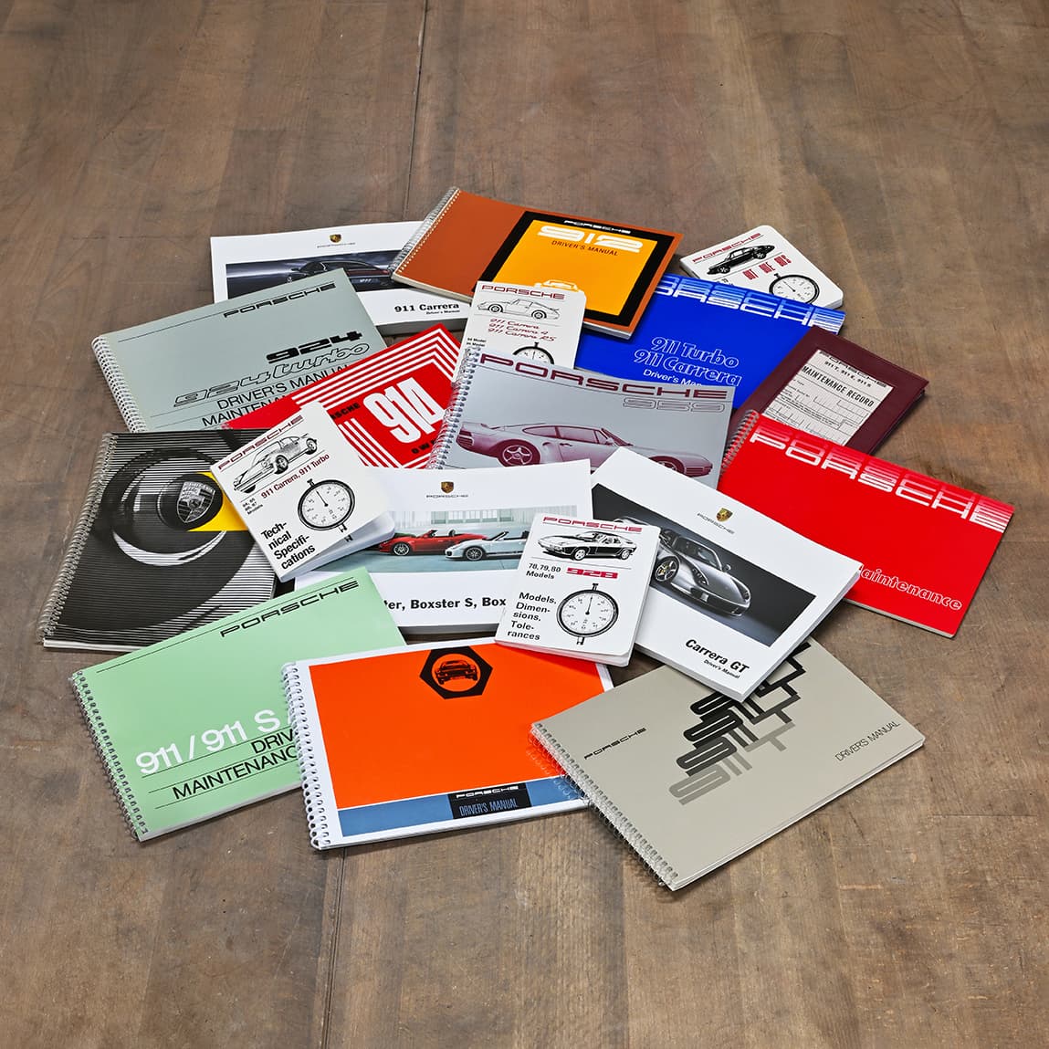 Sammlung originaler Porsche Betriebsanleitungen und technischer Literatur in verschiedenen Farben, ausgebreitet auf einem Holztisch.
