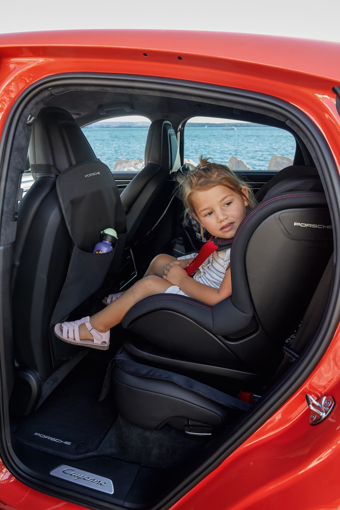 Enfant assis dans un siège enfant Porsche noir à l’arrière d’un Porsche Cayenne rouge, regardant vers l’extérieur, avec la mer en arrière-plan.