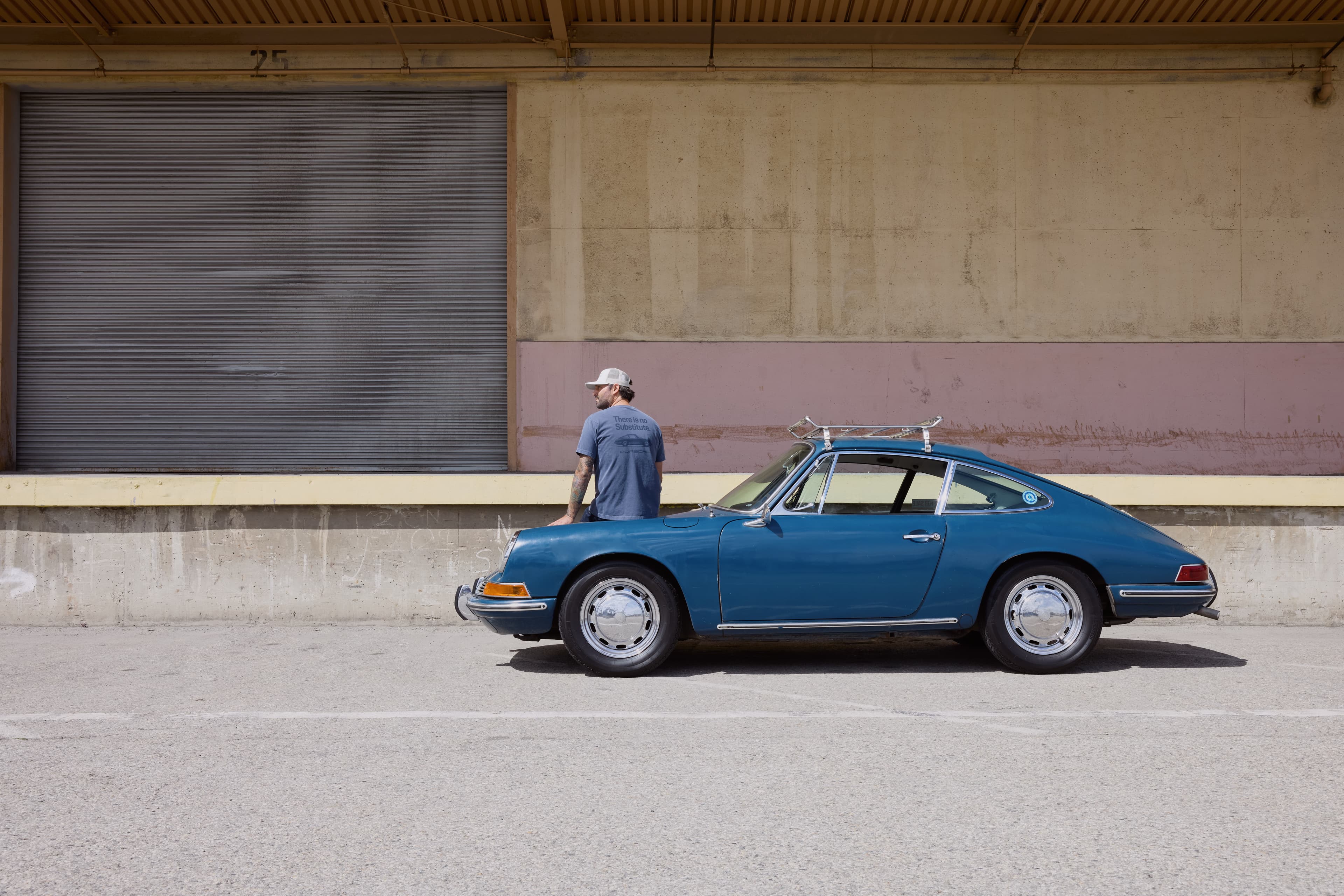 Man in a blue T-shirt next to a blue classic Porsche 911 with a roof rack in front of an industrial building.