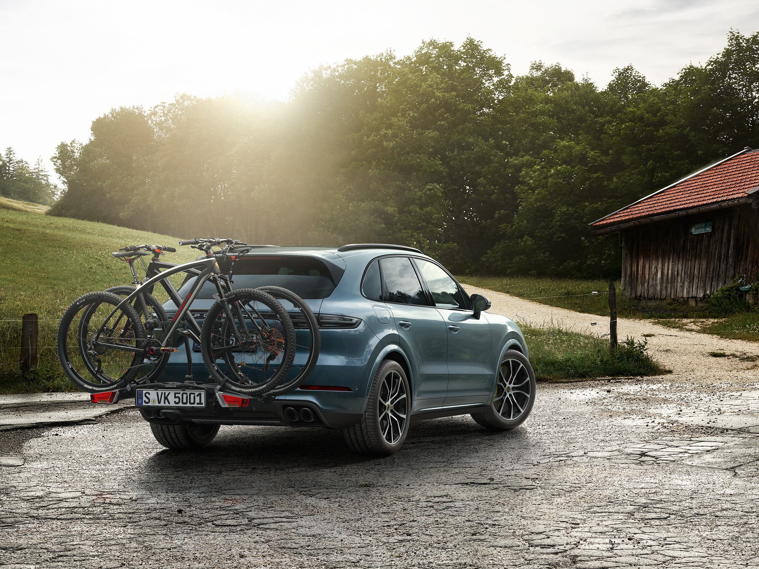 SUV Porsche azul com duas bicicletas de montanha num suporte traseiro para bicicletas, estacionado numa estrada rural ao pôr do sol.