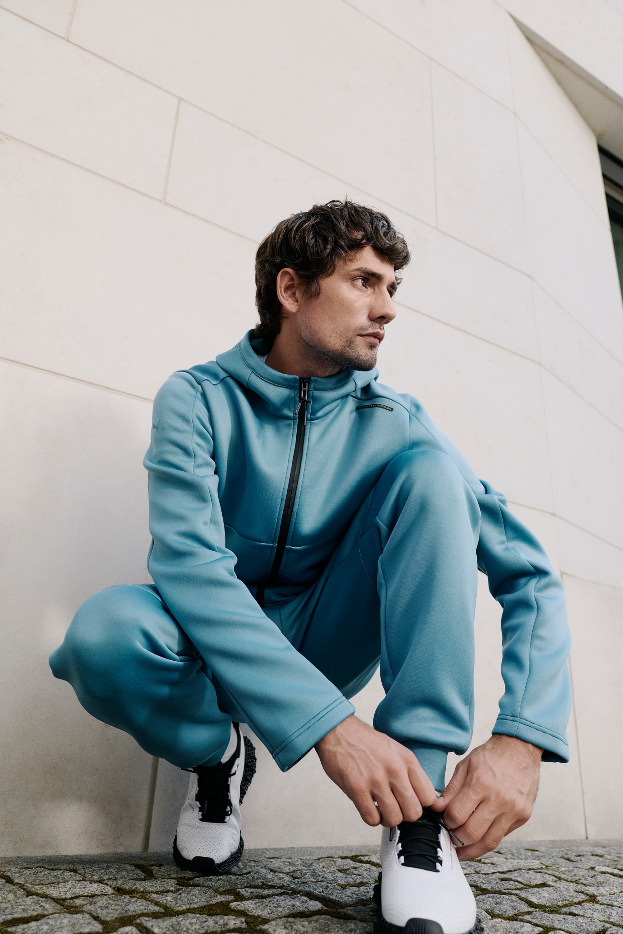 Man in blue tracksuit crouching, tying white sneakers, against a light stone wall.