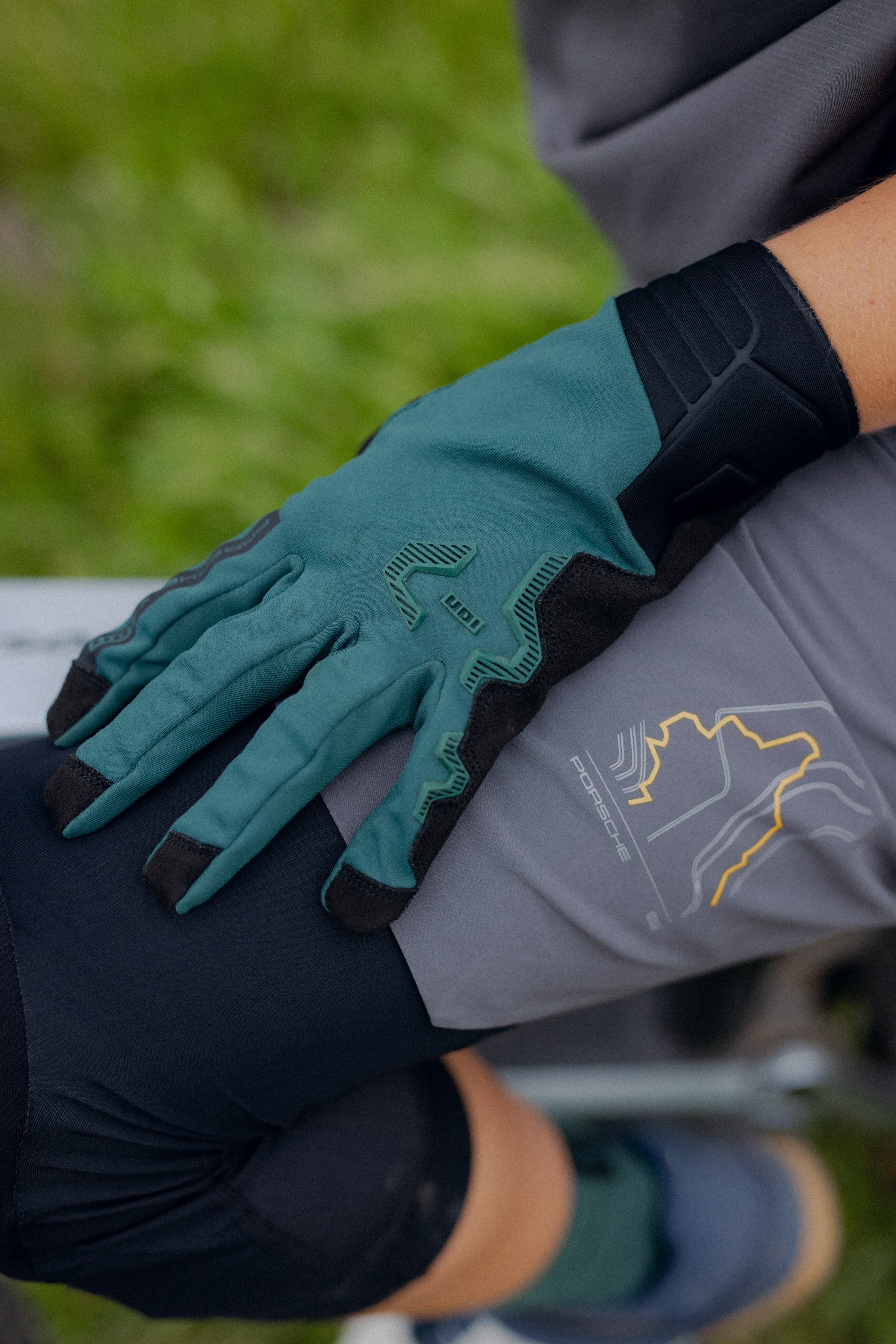 Turquoise cycling glove with black inserts on thigh, Porsche logo on grey shorts.