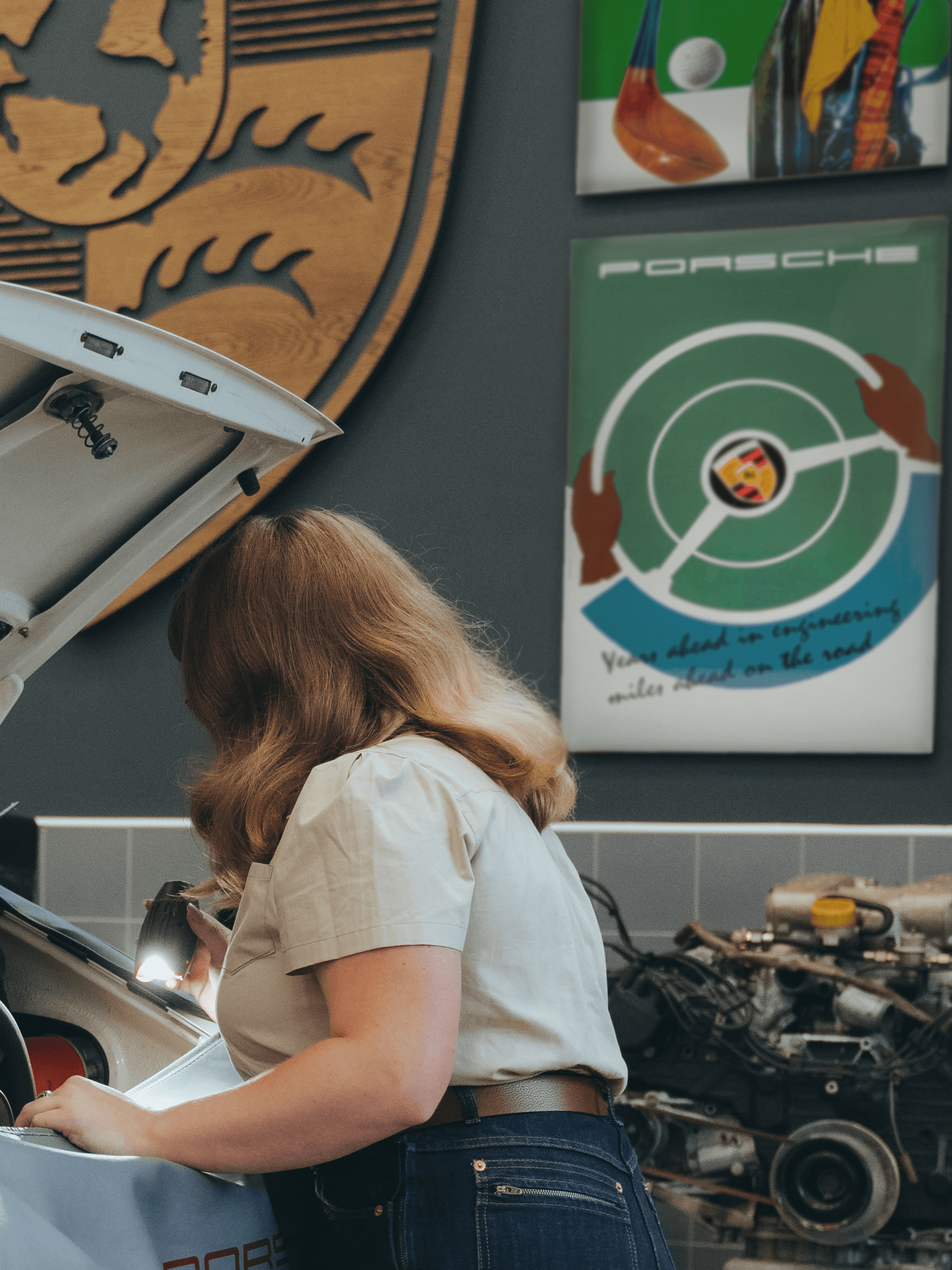 Woman is standing at the back of a car and in the background are different Porsche enamel plates on the wall