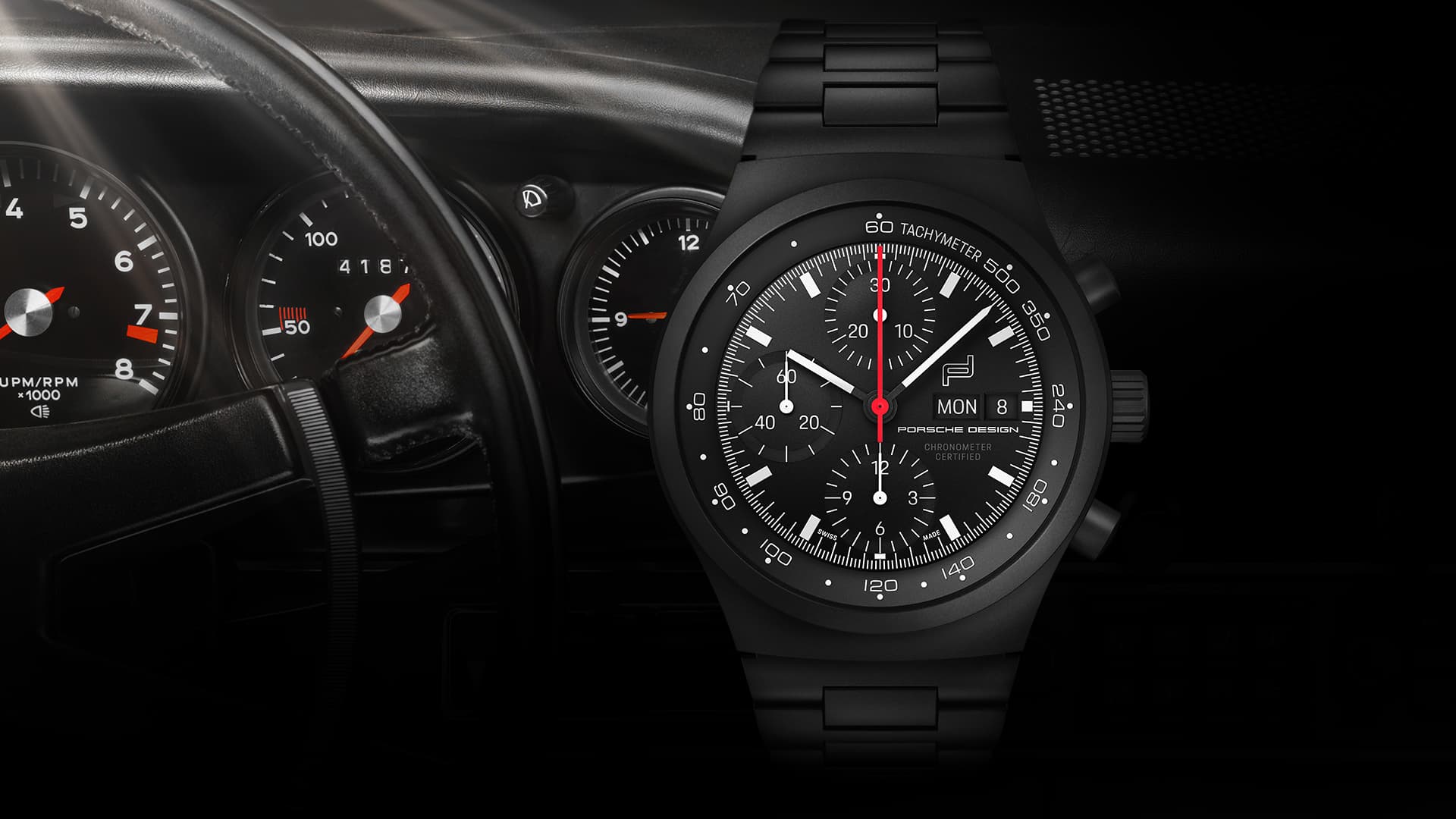 Black Porsche Design chronograph watch with red second hand, in front of a Porsche cockpit with instrument displays.