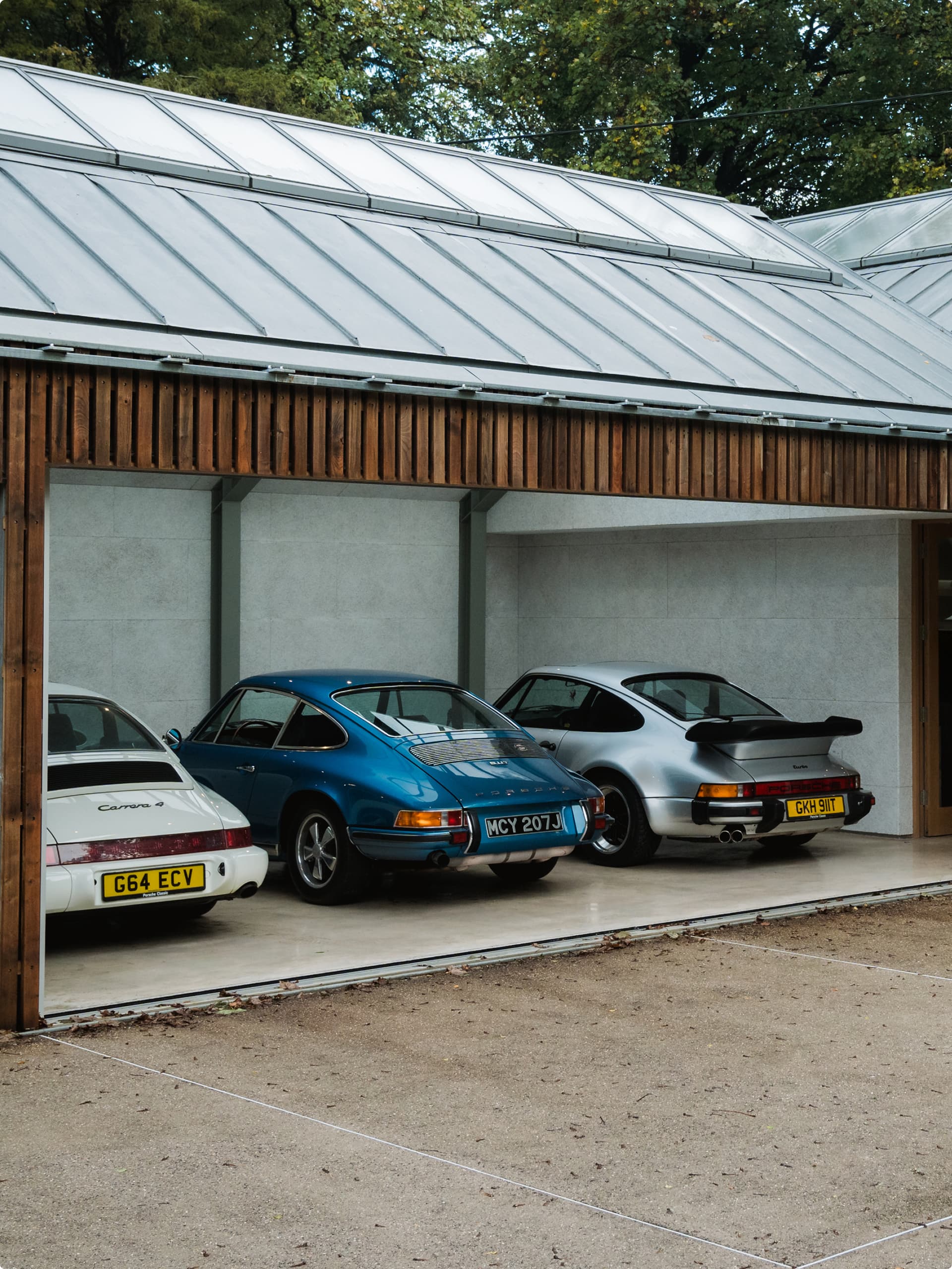 Three classic Porsche cars in white, blue, and silver parked in a modern garage with a wooden facade and glass roof.