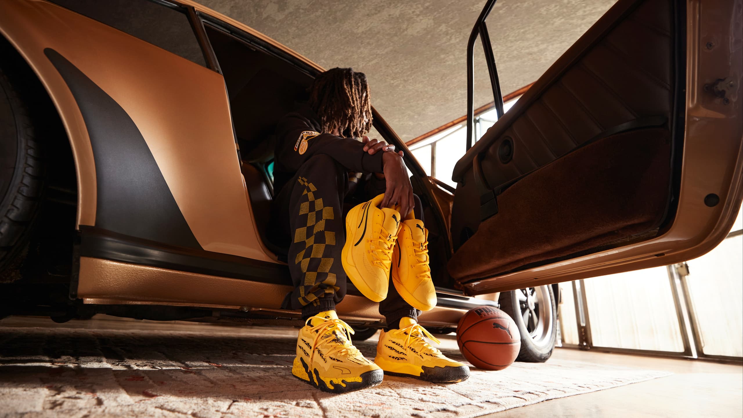 The image depicts a person sitting on the ground next to a tire and auto part. The individual is wearing footwear and appears to be indoors. The setting suggests a parked car nearby.