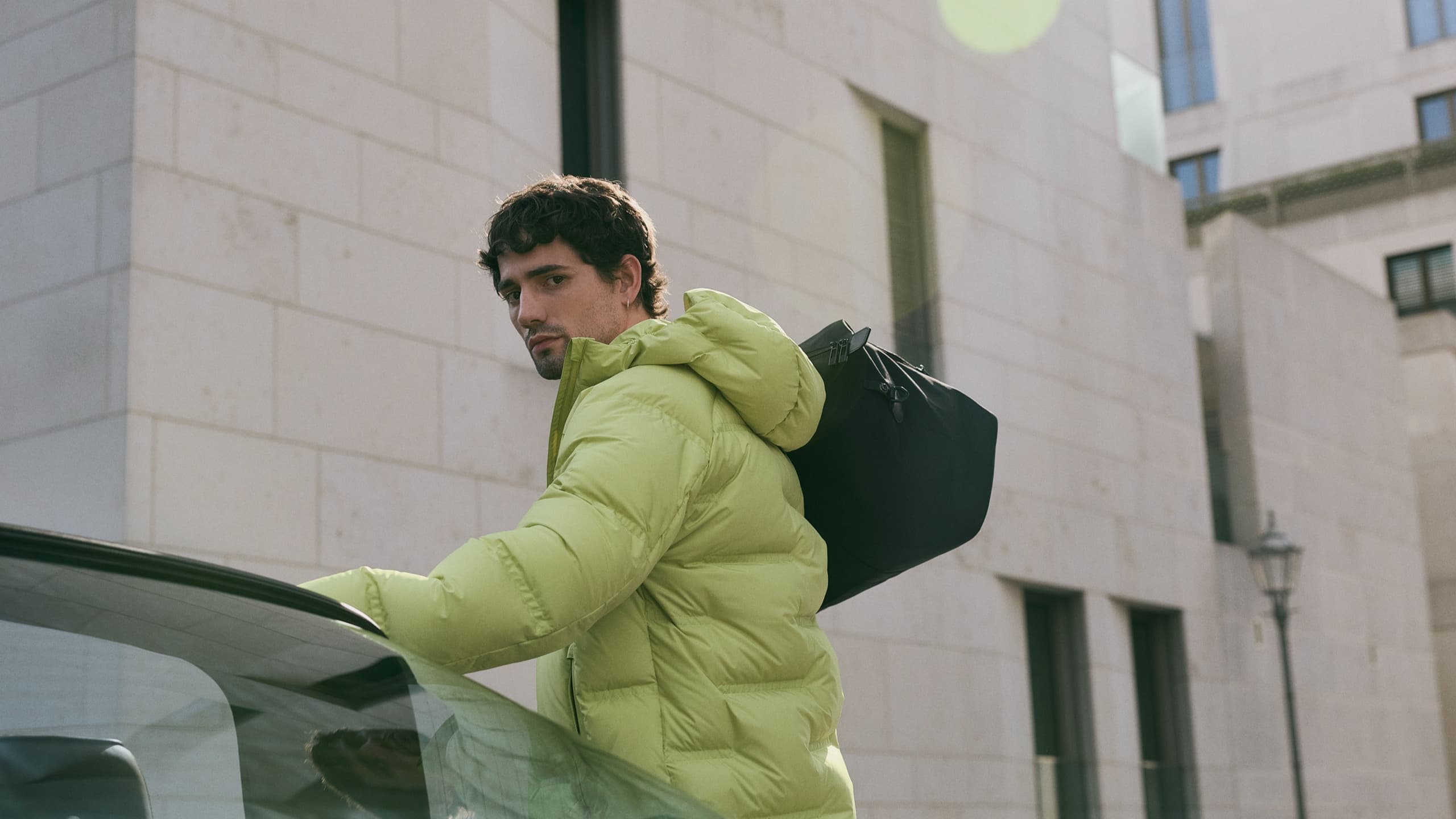 Man in lime green puffer jacket with black bag, standing by a car, against a modern stone building background.