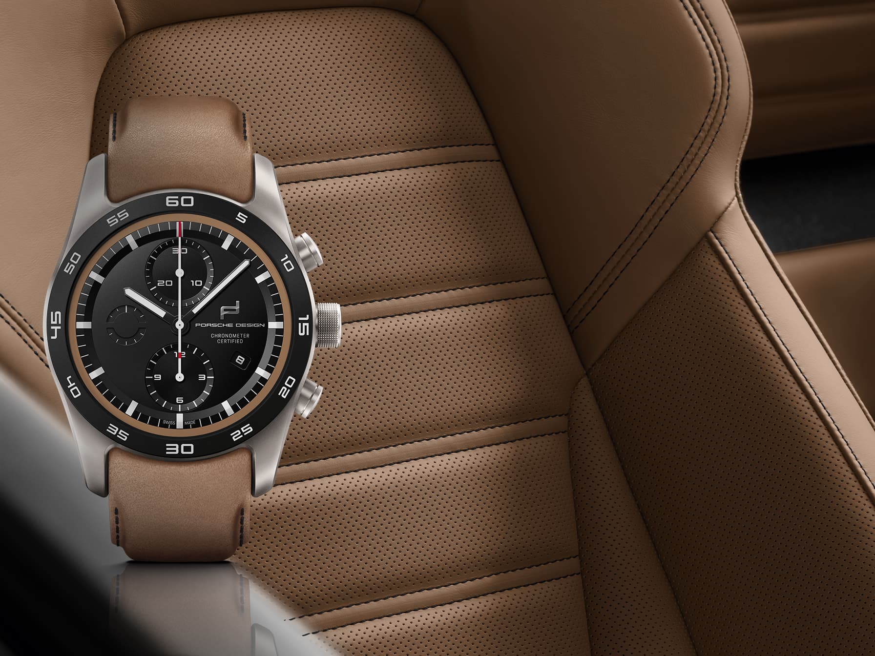 Chronographenuhr mit schwarzem Zifferblatt und braunem Lederarmband, vor einem lochperforierten, hellbraunen Ledersitz im Auto.
