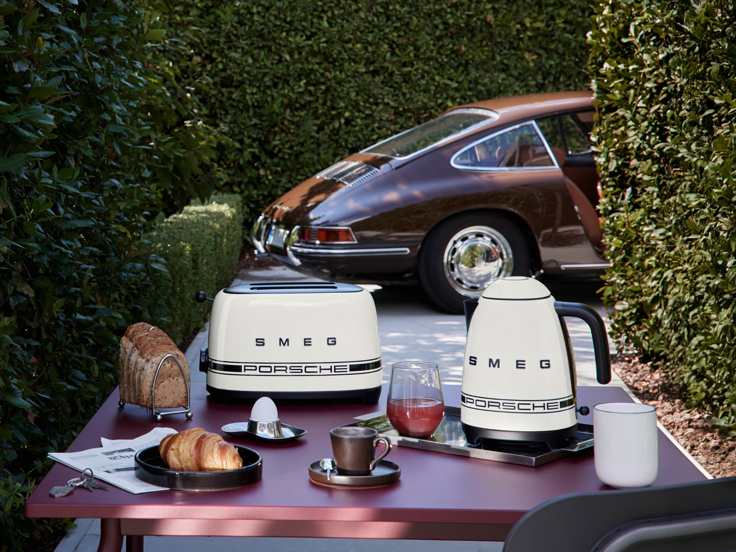 Porsche x Smeg kettle and toaster in Carrara White on an outdoor breakfast table, Porsche 911 in the background