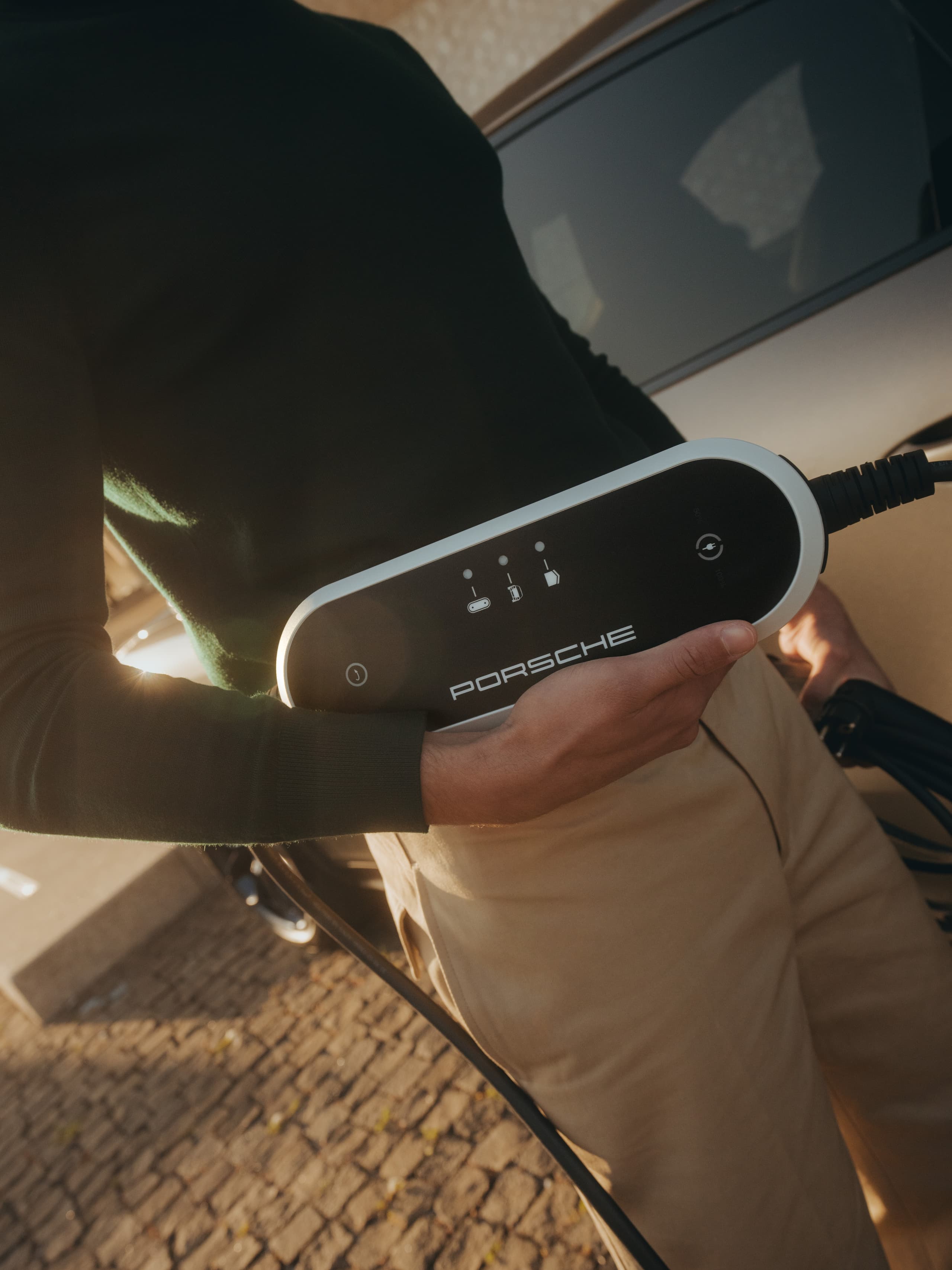 Personne tenant un chargeur de voiture électrique Porsche, portant un pull foncé et un pantalon beige, se tenant près d'une voiture sur une rue pavée.