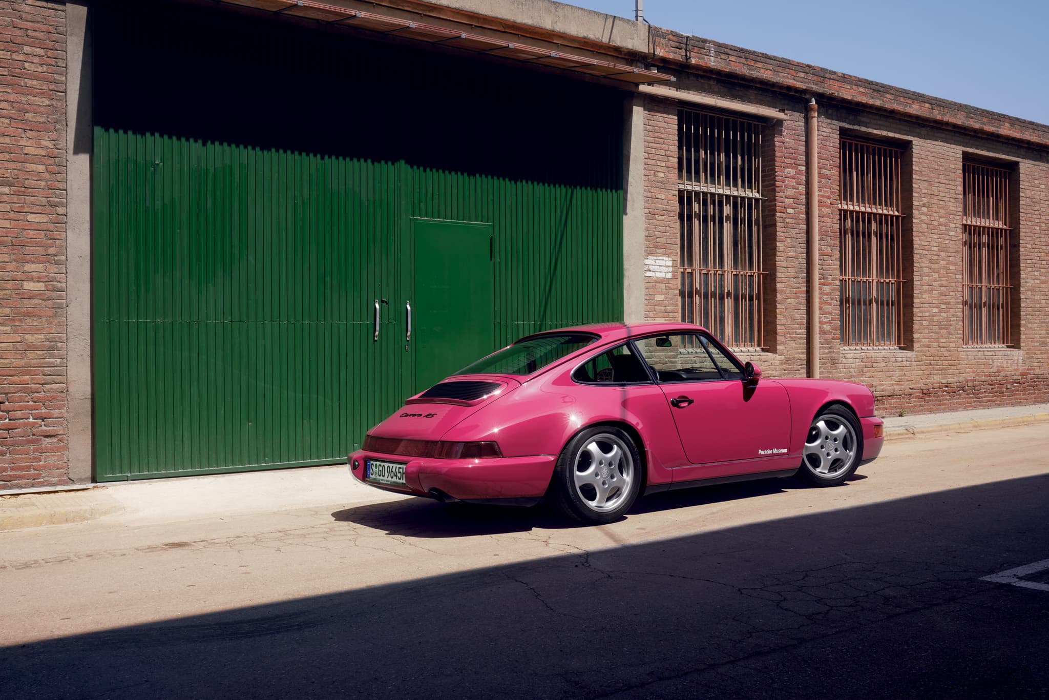 A pink historic Porsche car in front of a garage with green doors