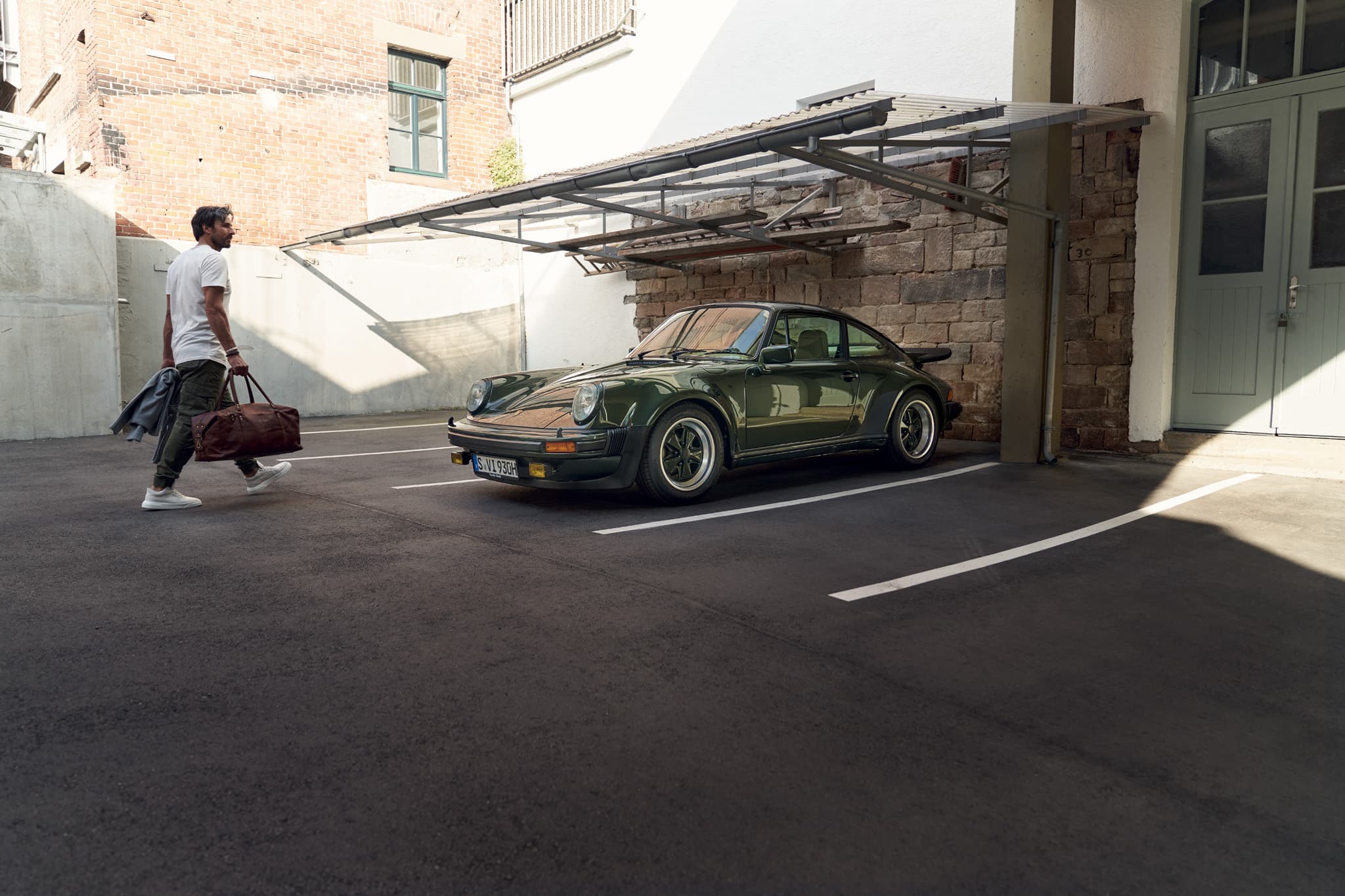 Man carrying a travel bag walks toward a dark green Porsche 911 parked under a carport in a courtyard.