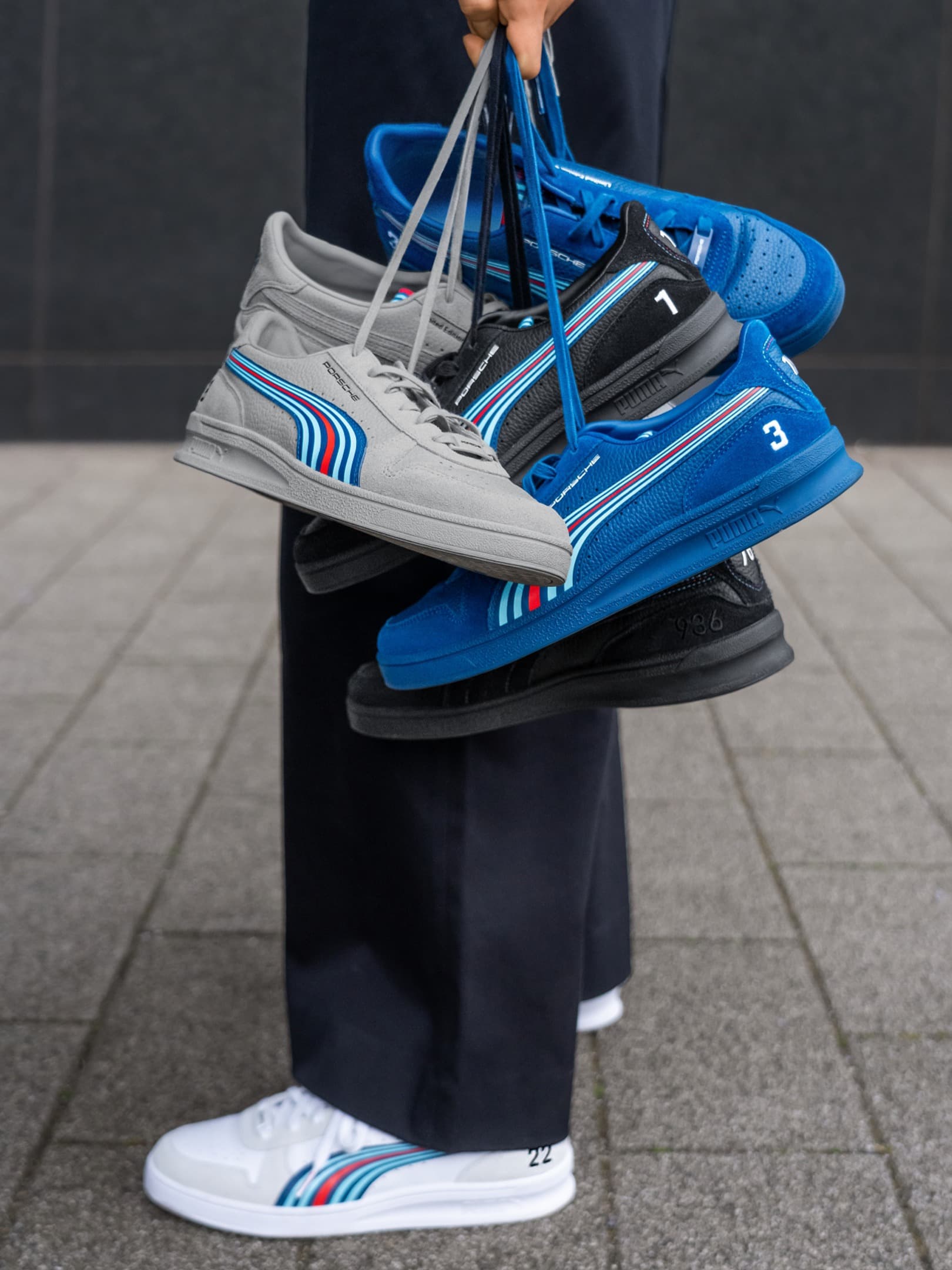 Person holding gray, black, and blue sneakers with colorful stripes, standing on a tiled surface.