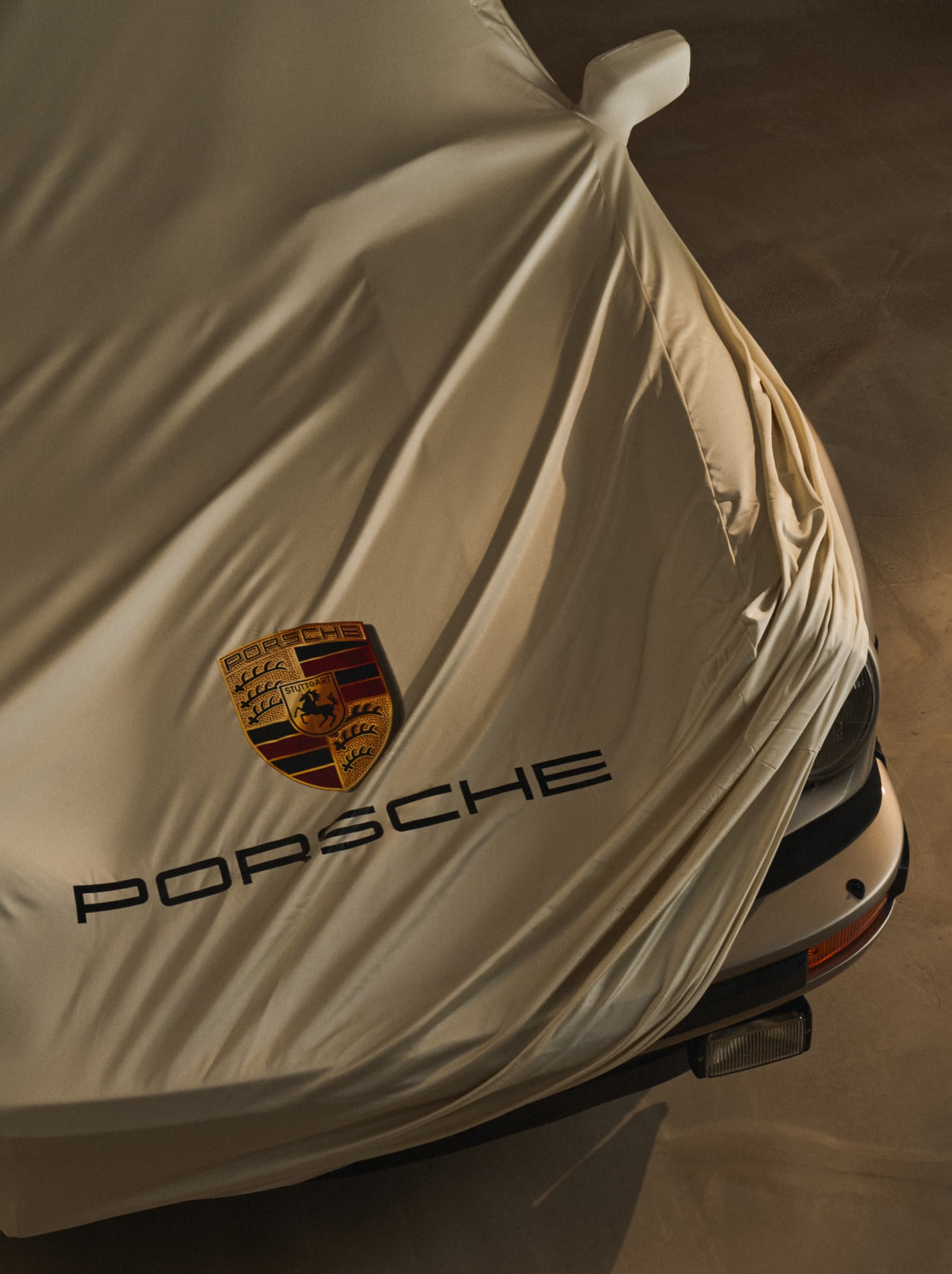 Car covered with beige fabric, displaying Porsche logo and text, partially revealing the front of the vehicle.