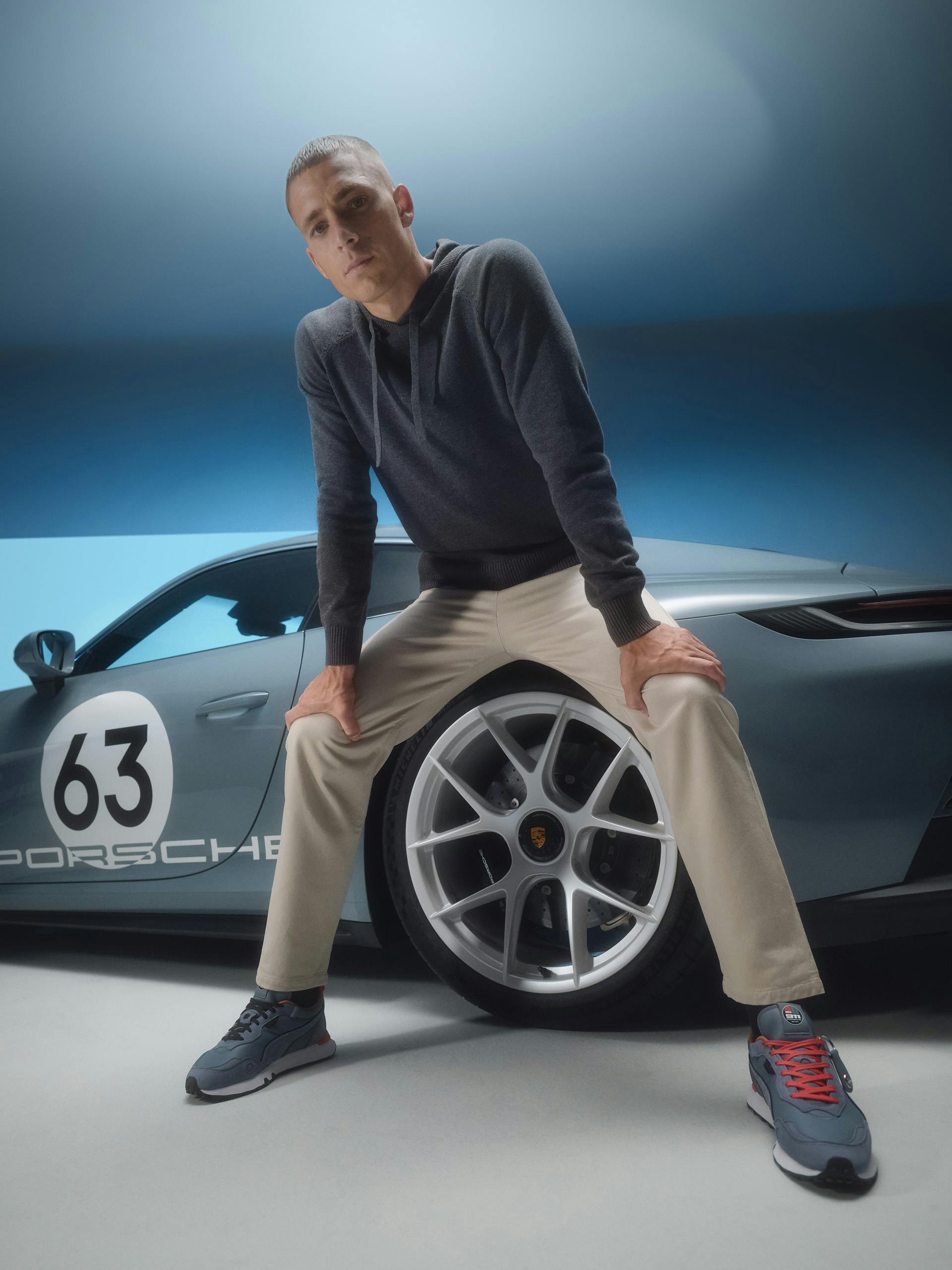 A man in brown trousers, a dark blue sweatshirt and blue Puma sneakers can be seen sitting on the tyre of the 911 Carrera S/T, looking head-on into the camera.