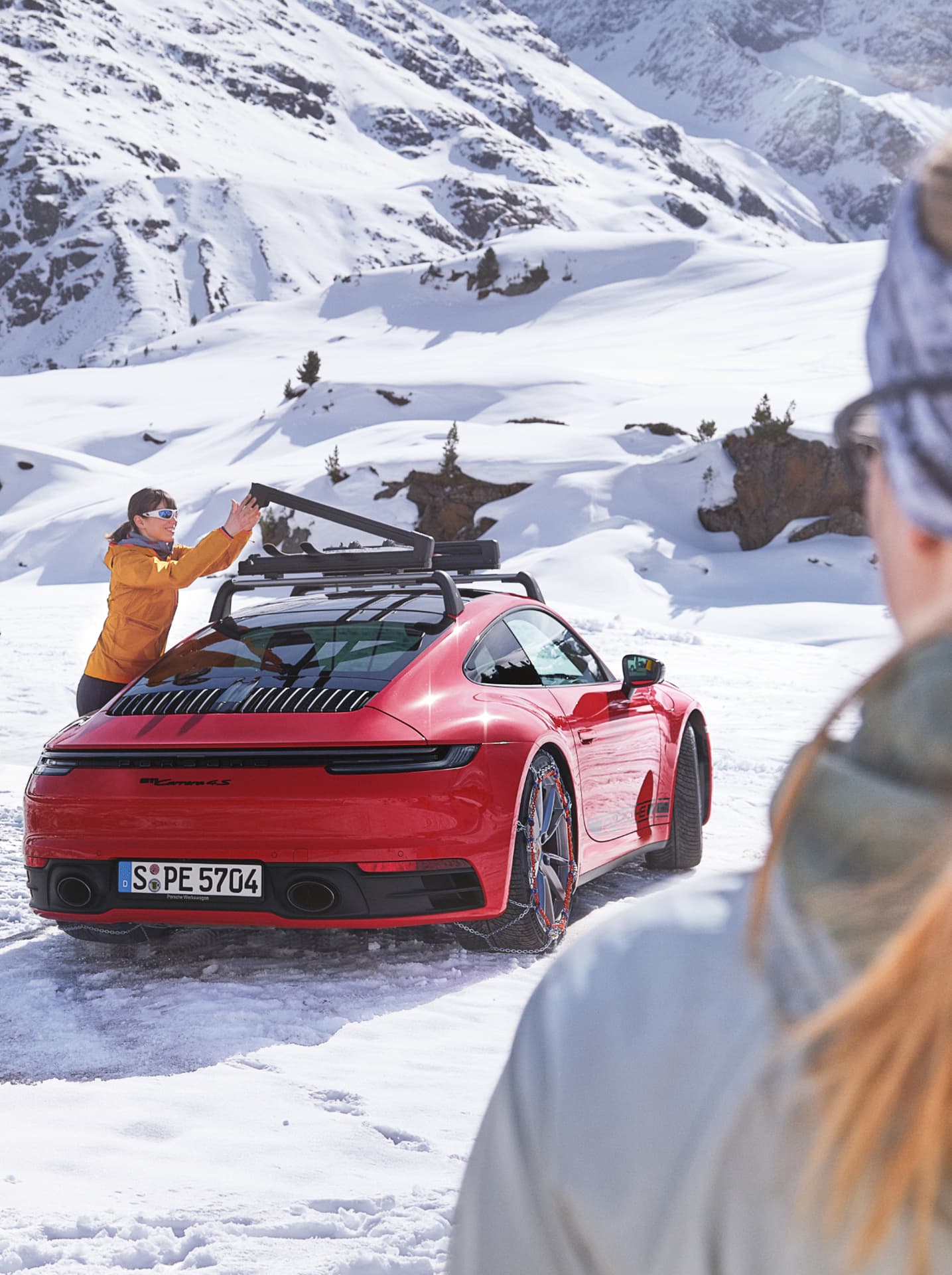 Red Porsche 911 with snow chains, person in orange jacket adjusts roof rack, snowy mountain backdrop.