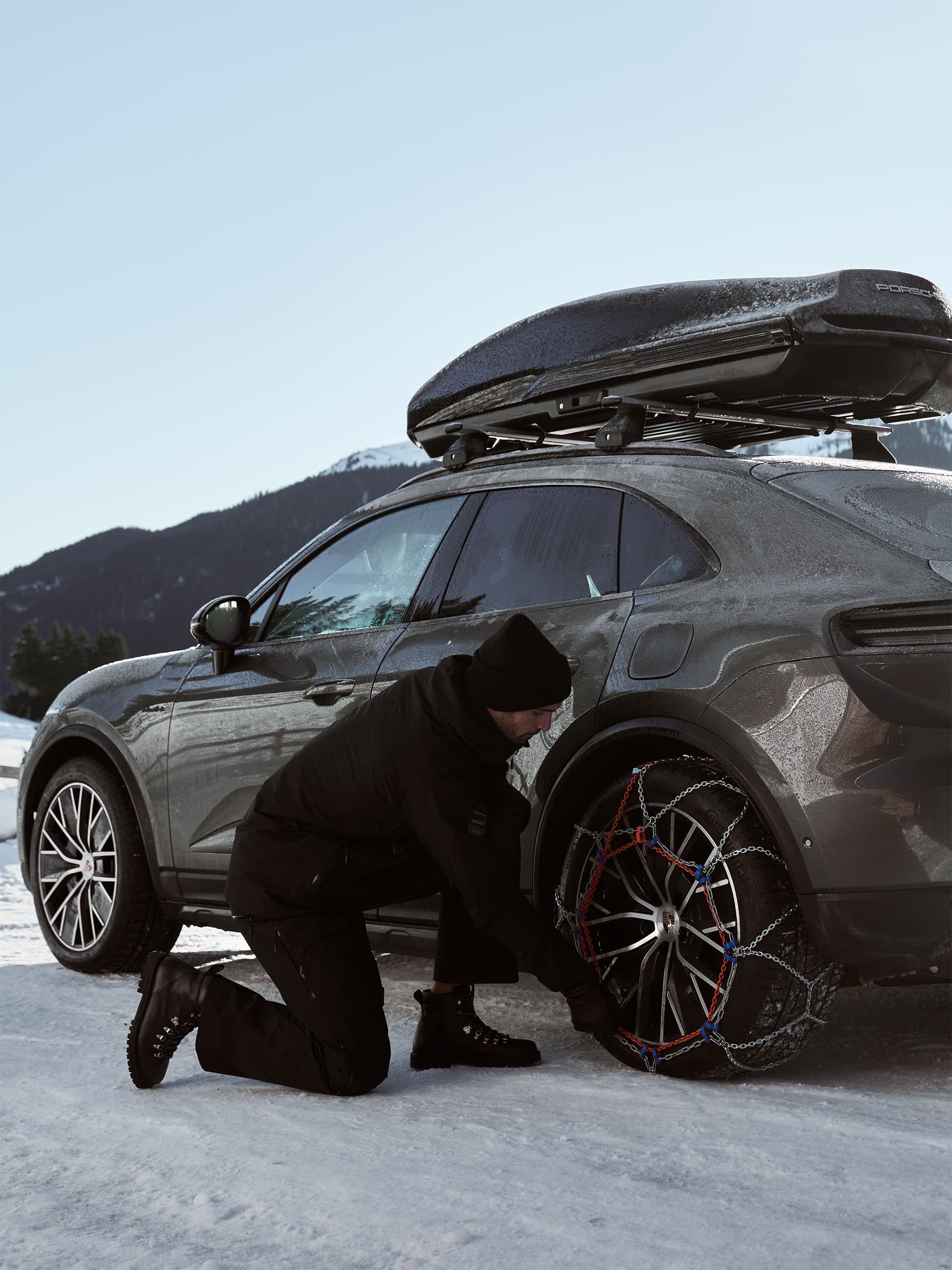 Person fitting snow chains on a gray Porsche with roof box in snowy mountain setting.