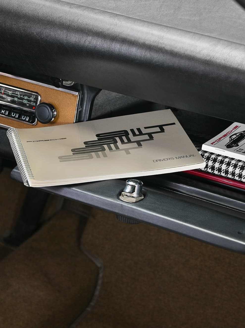 Porsche driver's manual in a car glove compartment, next to a houndstooth-patterned book, with a vintage radio visible.