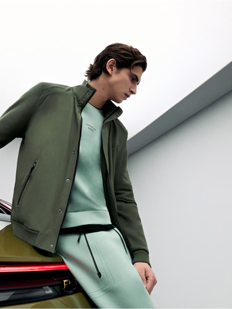 Homme en veste verte et sweat-shirt menthe s'appuyant sur une voiture, sur un fond minimaliste blanc et gris.