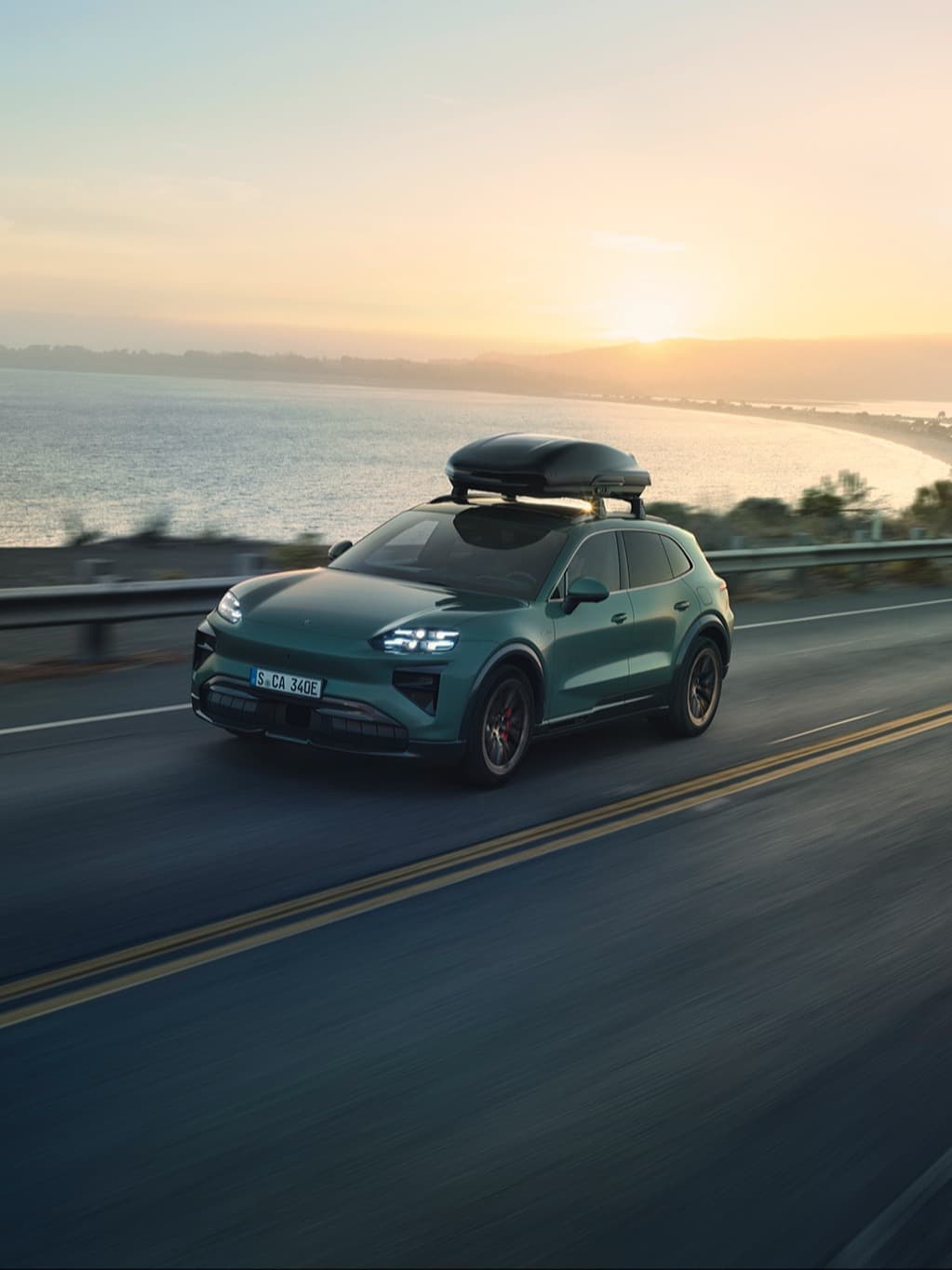 Green Porsche SUV with roof box driving on coastal road at sunset, ocean view in background.
