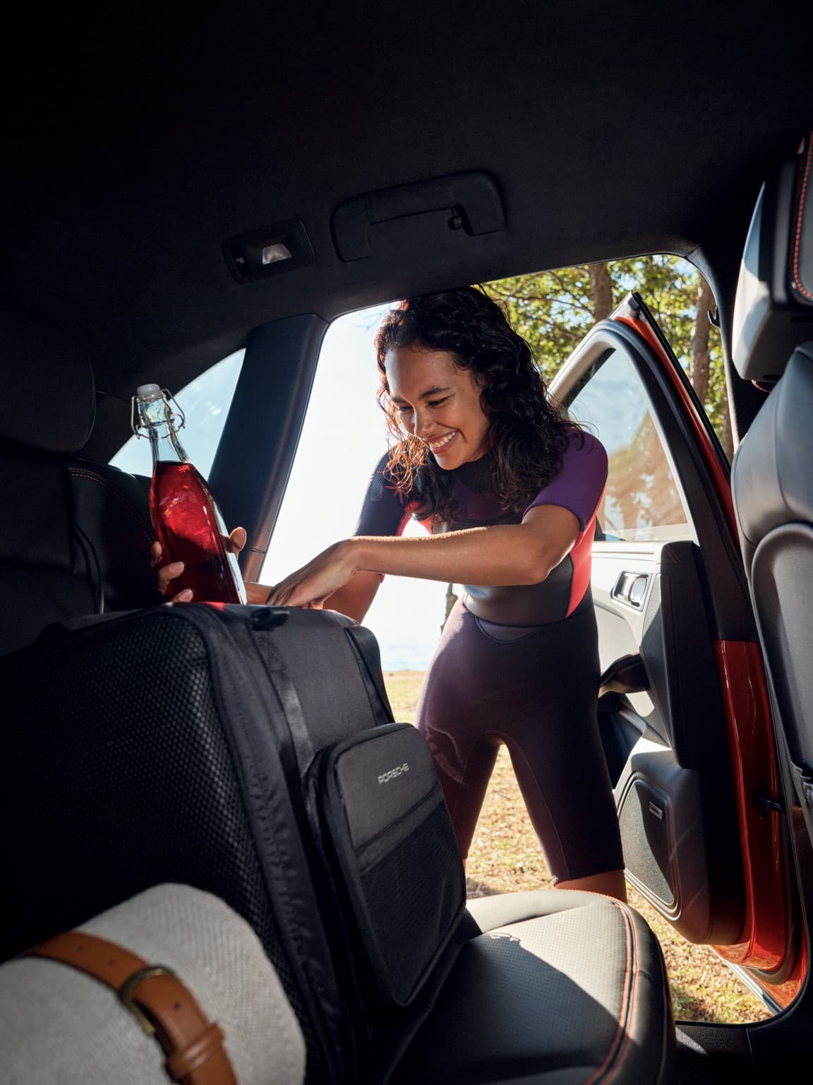 Woman loads a black Porsche seatback bag into the rear passenger area next to a red glass bottle, car door open outdoors.