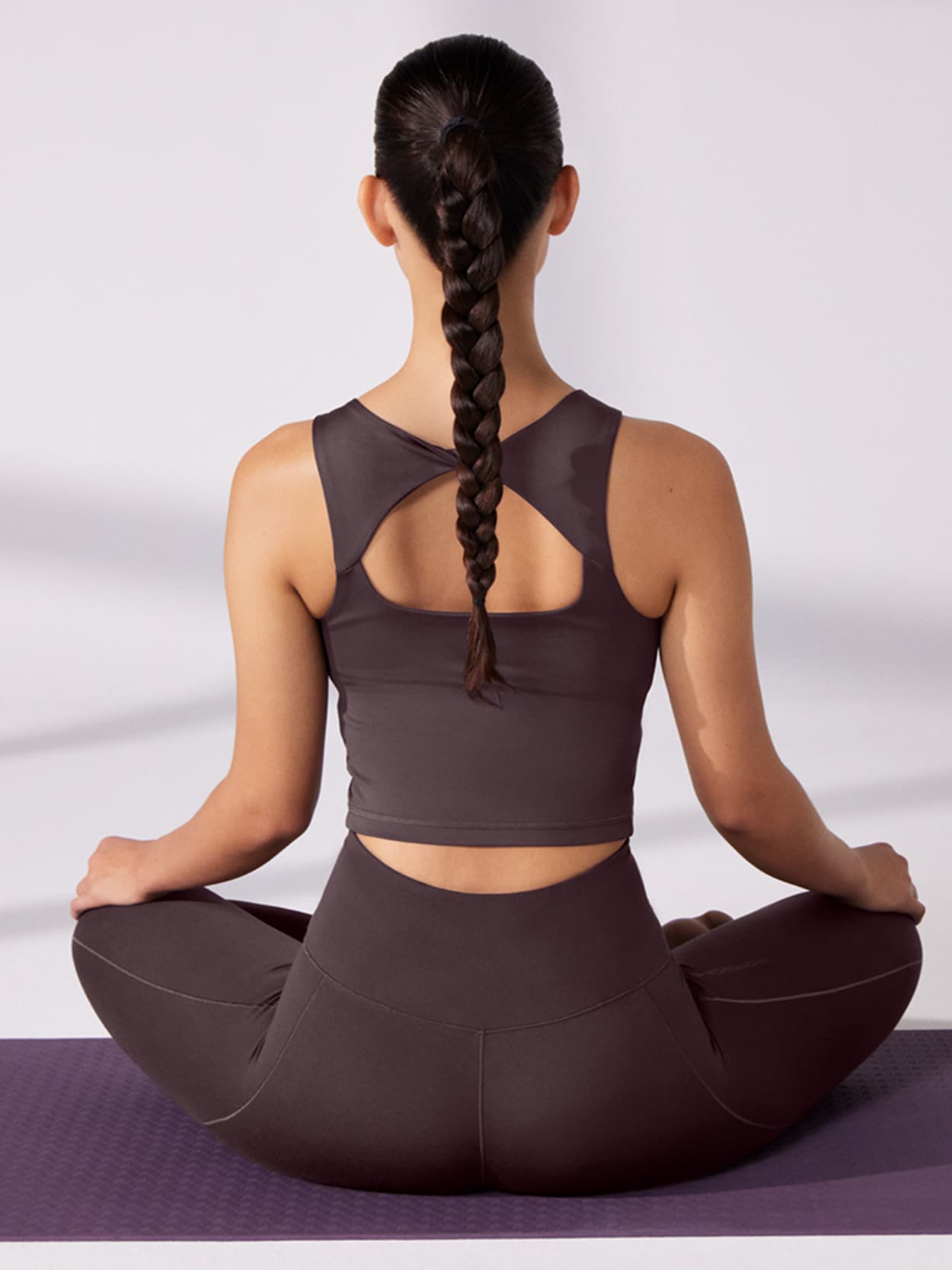 Woman in dark sportswear sitting cross-legged on a purple yoga mat, hair in a long braid, viewed from behind.