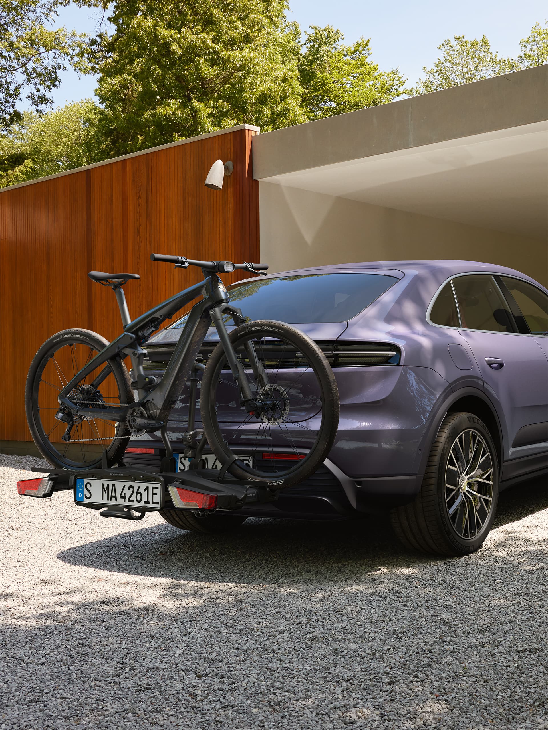 Fioletowy SUV Porsche z tylnym bagażnikiem rowerowym i zamontowanym czarnym rowerem elektrycznym, zaparkowany przed nowoczesnym garażem wśród zieleni.