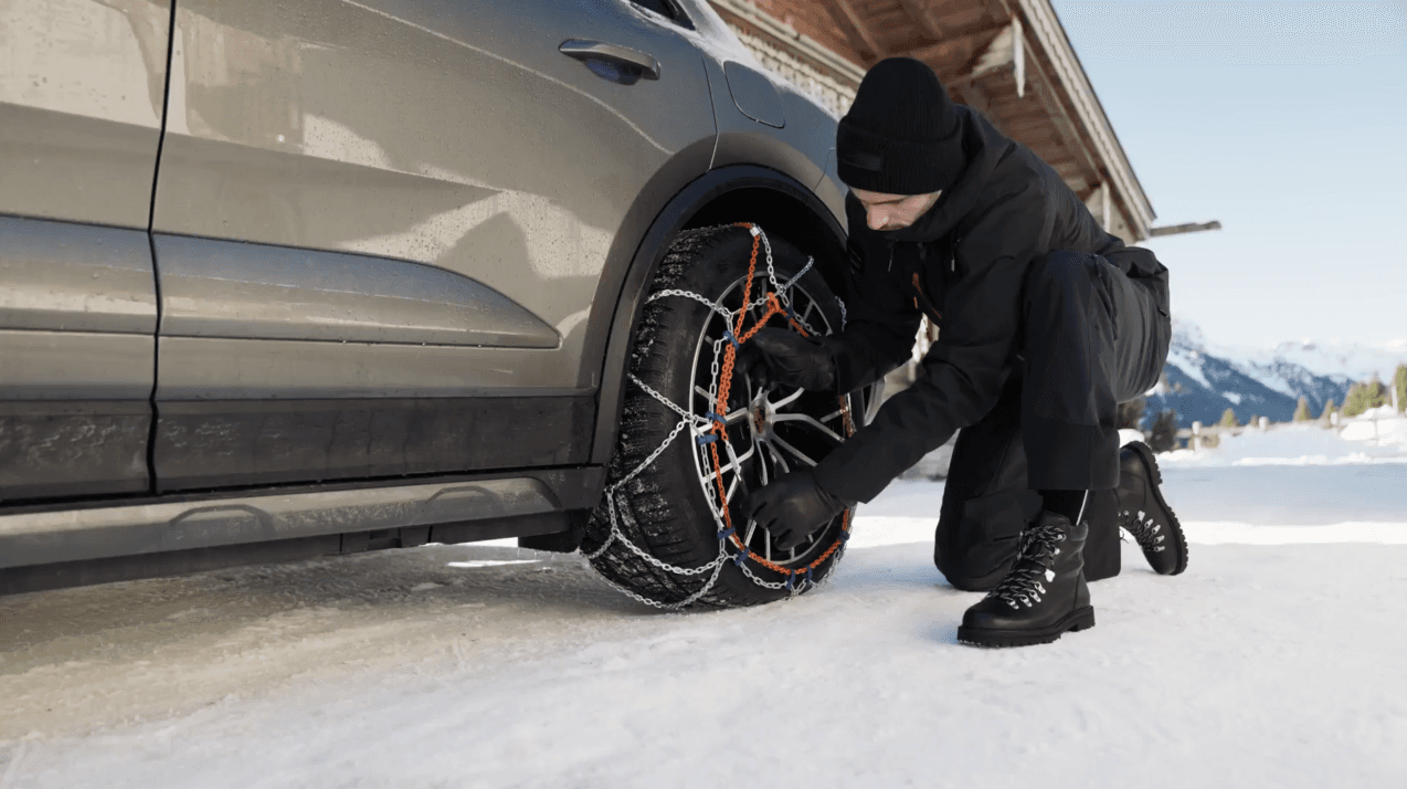 Person fitting snow chains on a car tire in a snowy mountain setting, wearing black winter clothing and boots