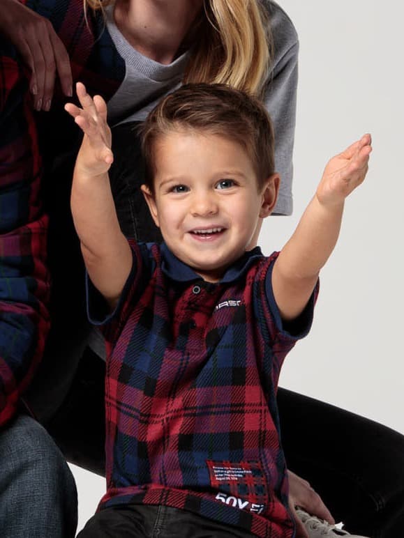 Smiling child in a red and blue plaid shirt with arms raised, sitting in front of an adult on a light background.