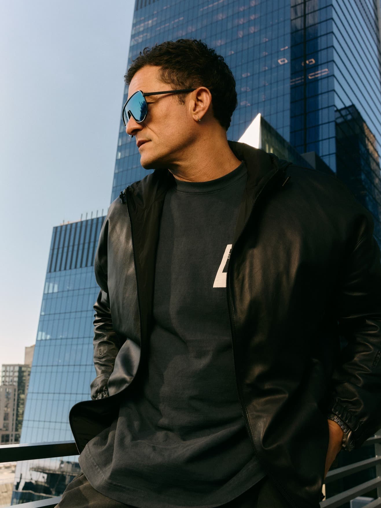 Homme portant des lunettes de soleil miroir et une veste en cuir noir, devant des gratte-ciel en verre en ville.