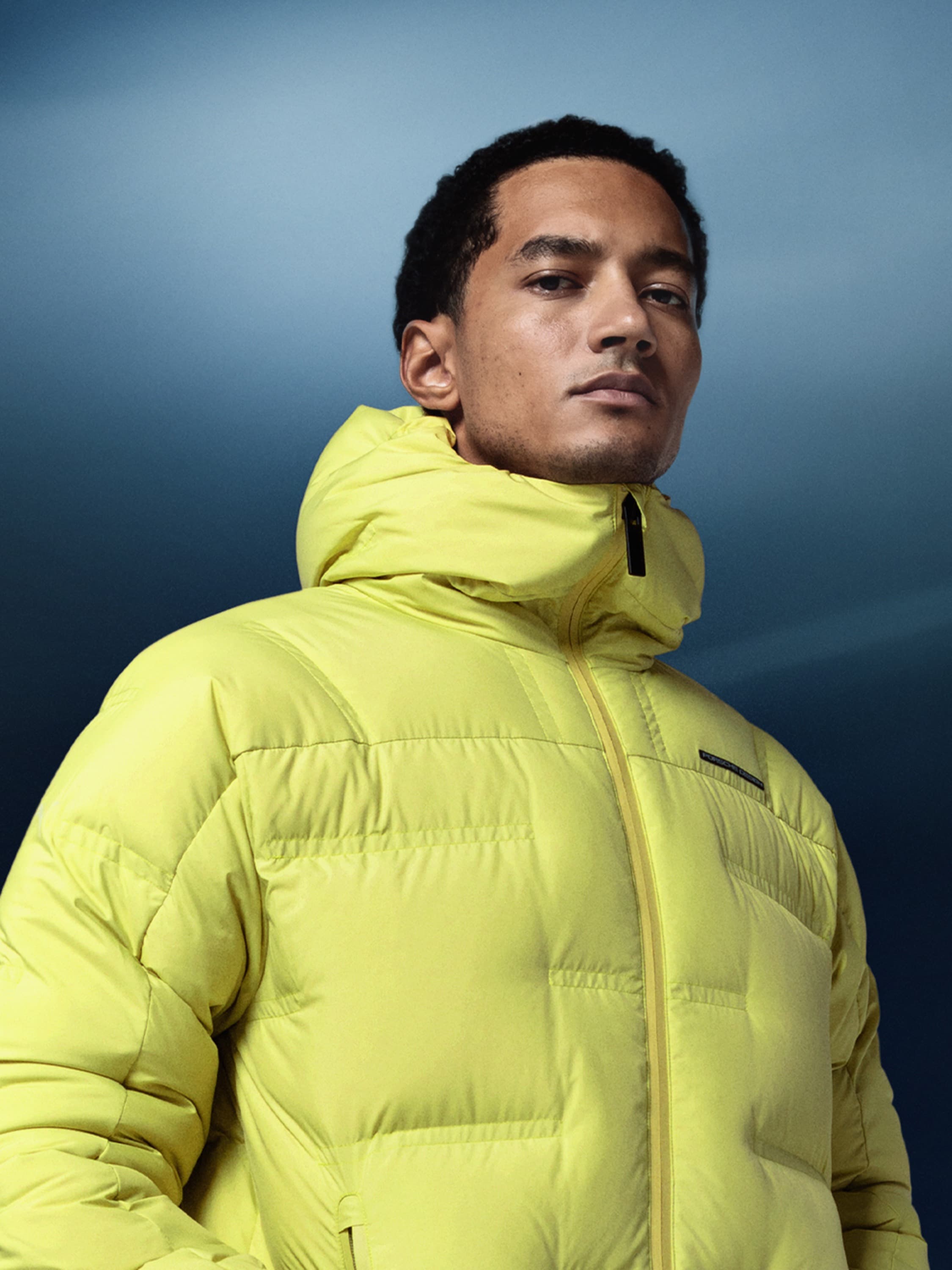 Mann in einer leuchtend gelben Daunenjacke mit Kapuze vor einem blauen Hintergrund.