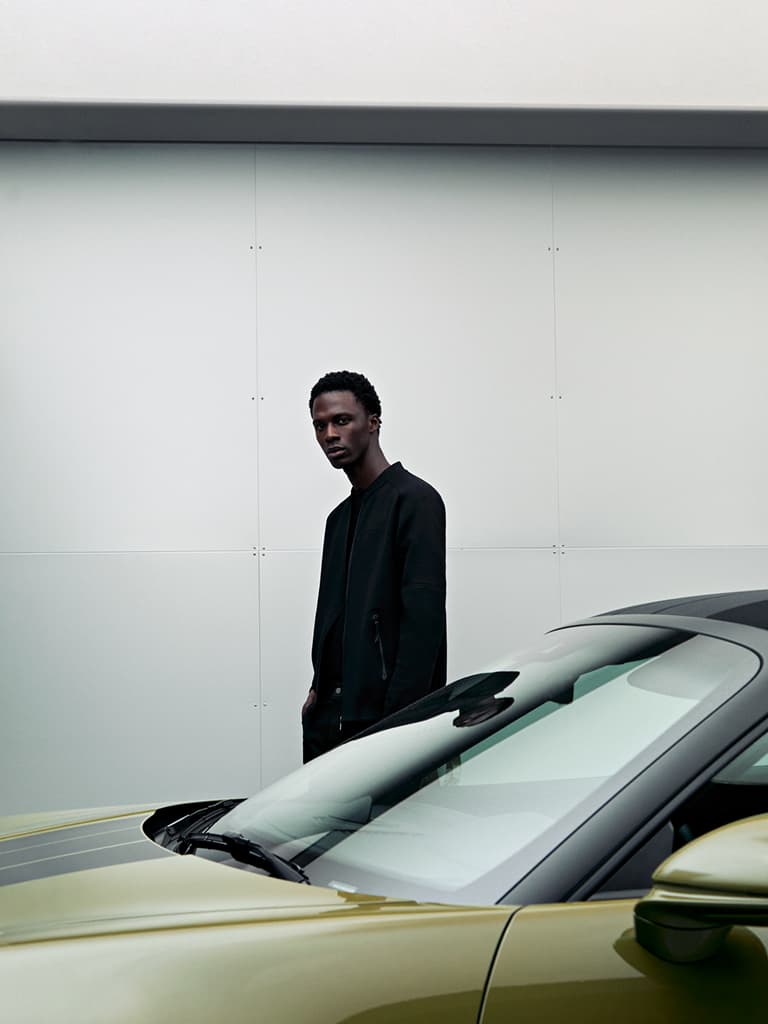 Mann in schwarzer Jacke steht neben einem eleganten, grünen Auto vor einer minimalistischen weißen Wand.