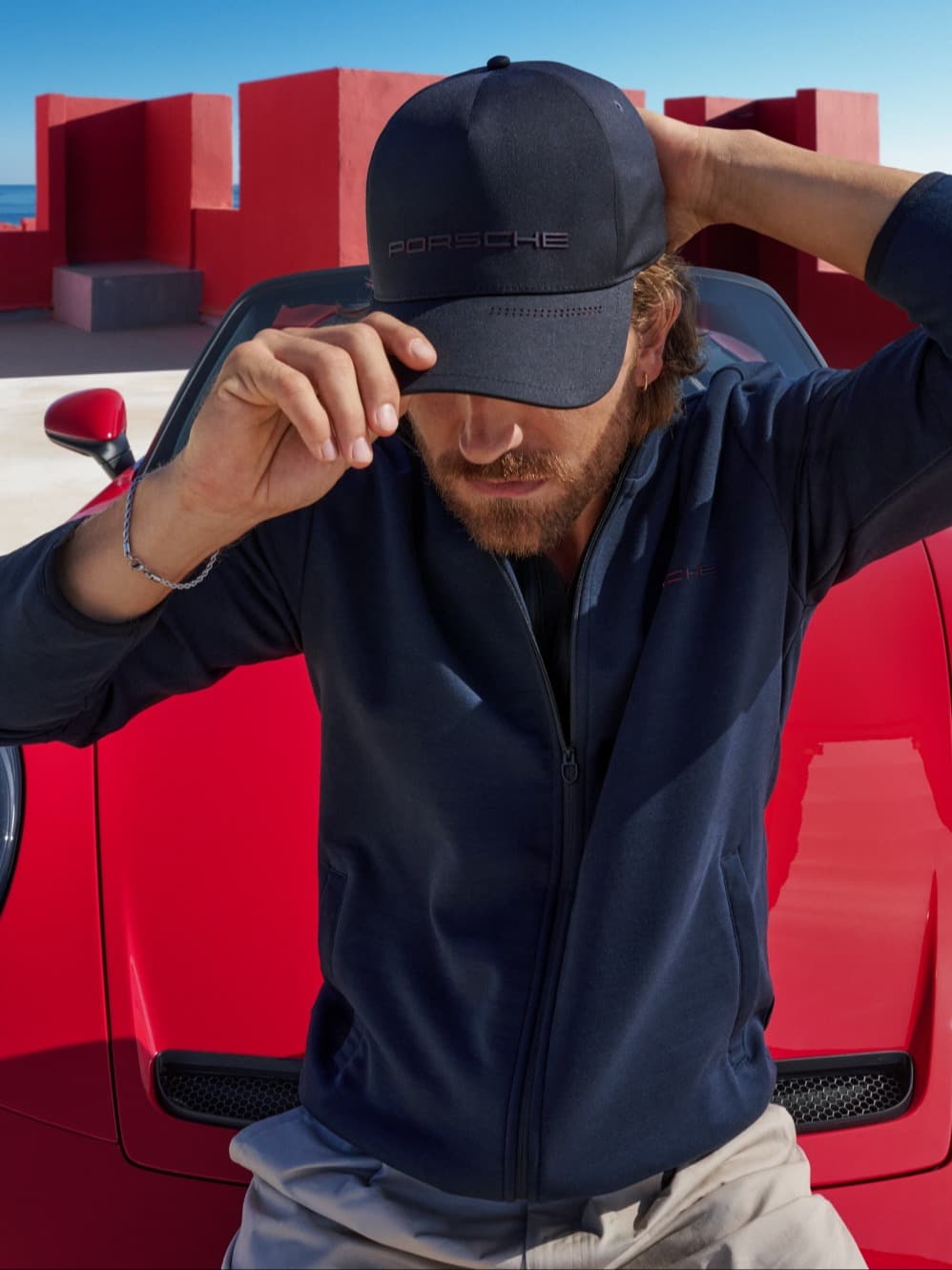 Man sits on the hood of a red 911 S/C holding his 911 S/C Collection cap.